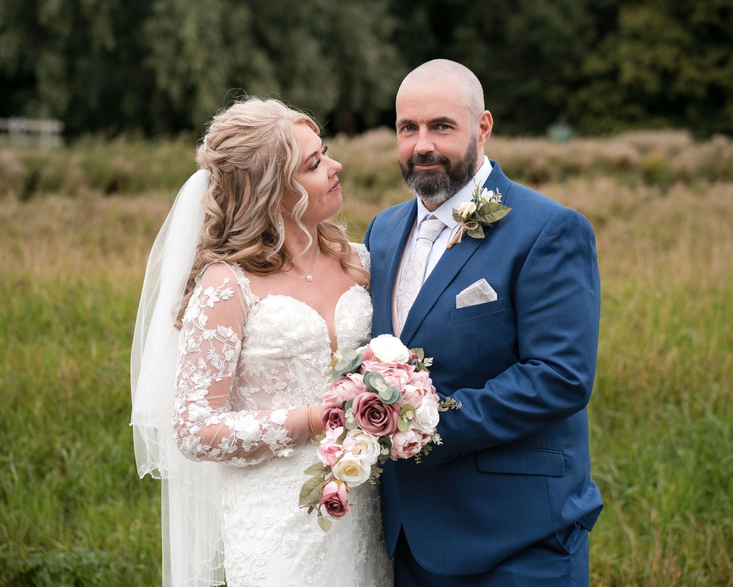 Newlyweds share a tender moment, bride holding floral bouquet in lace dress, groom in navy suit with boutonniere.