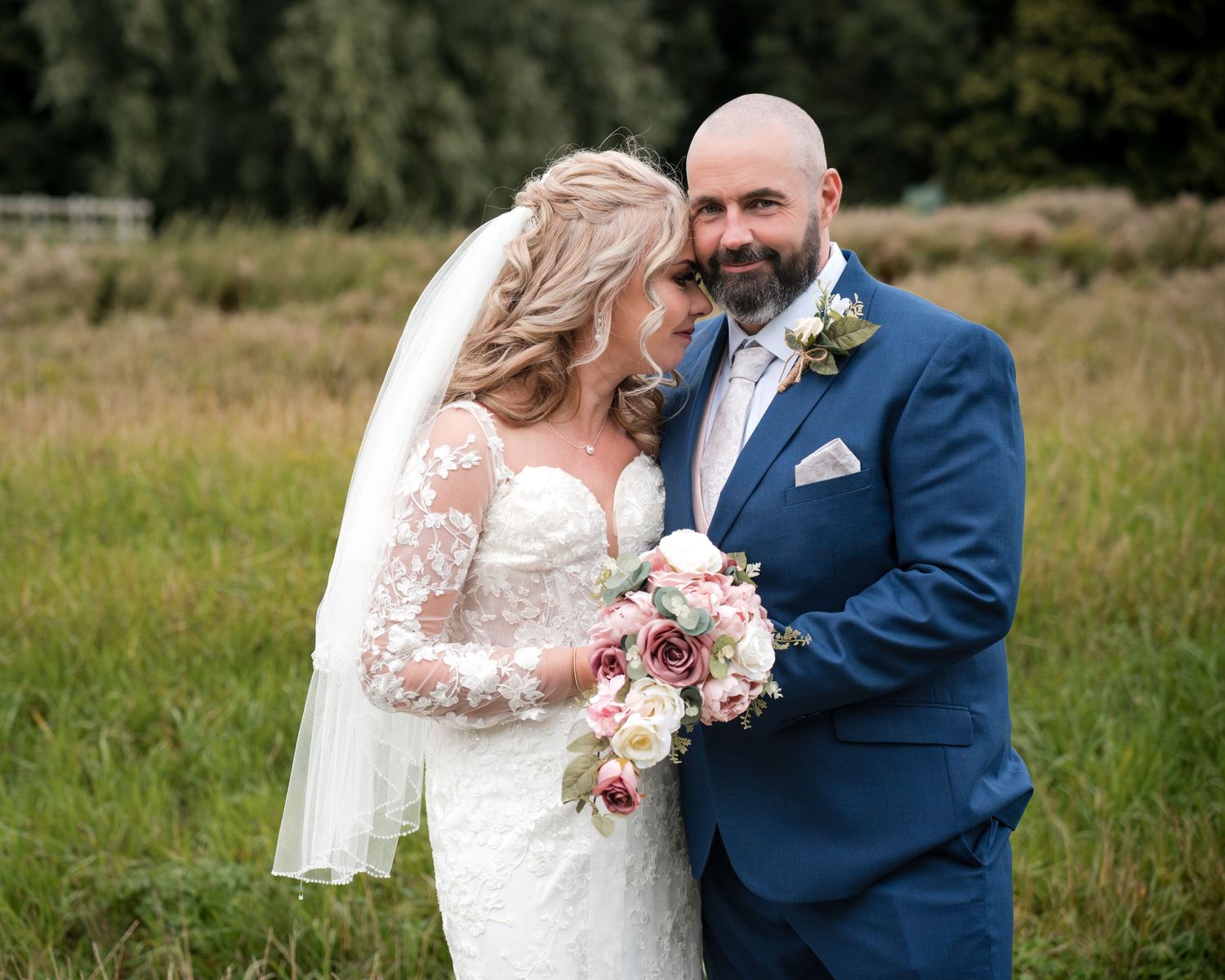 Bride and groom posing outdoors on wedding day, bride in lace dress with veil holding bouquet, groom in navy suit.