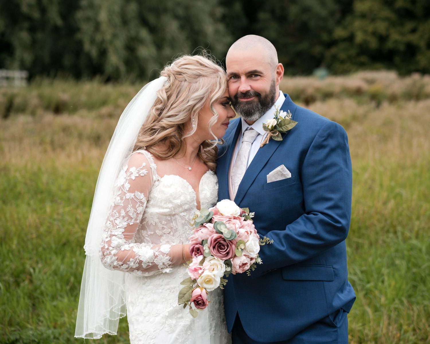 Bride and groom share romantic moment outdoors, she holds pink bouquet in lace gown, he wears navy suit with boutonniere.