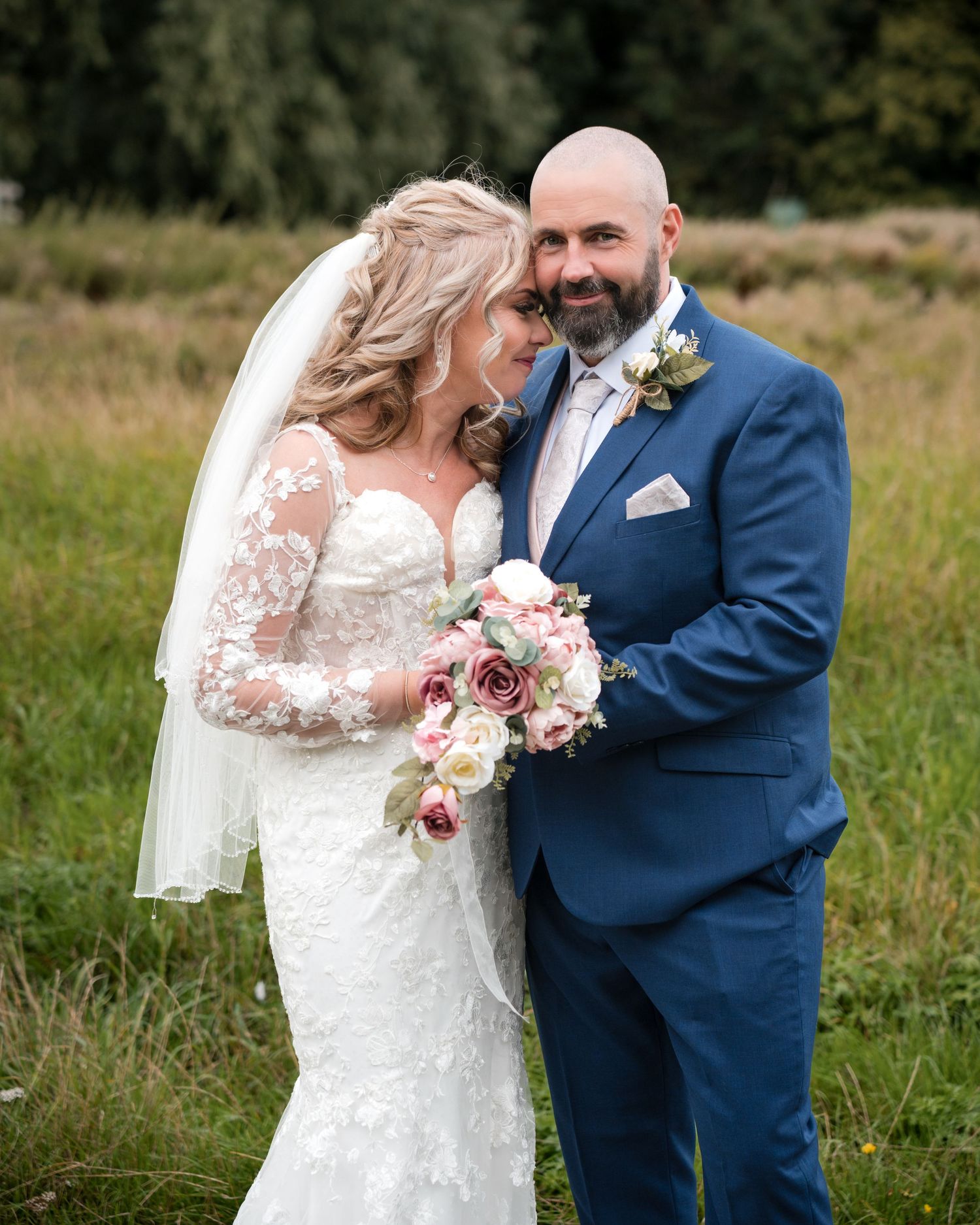 Bride in lace gown with bouquet and groom in blue suit share romantic moment in meadow.