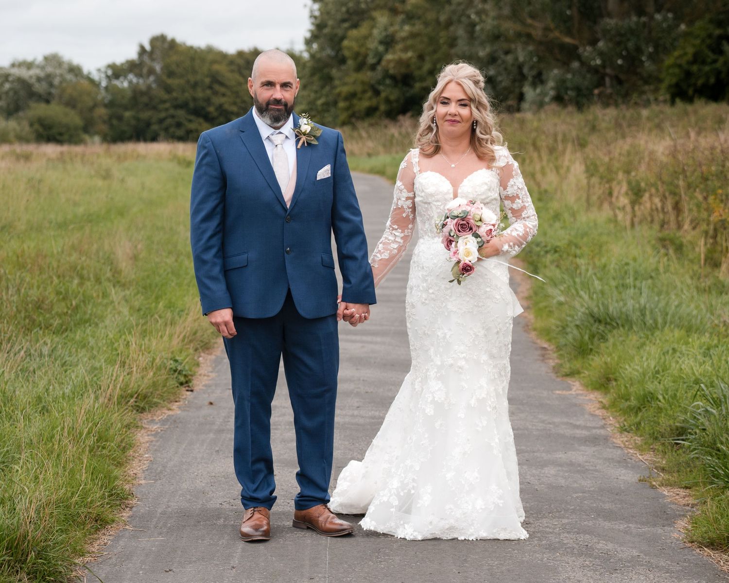 Bride in lace gown and groom in blue suit hold hands on a path surrounded by green fields on their wedding day.