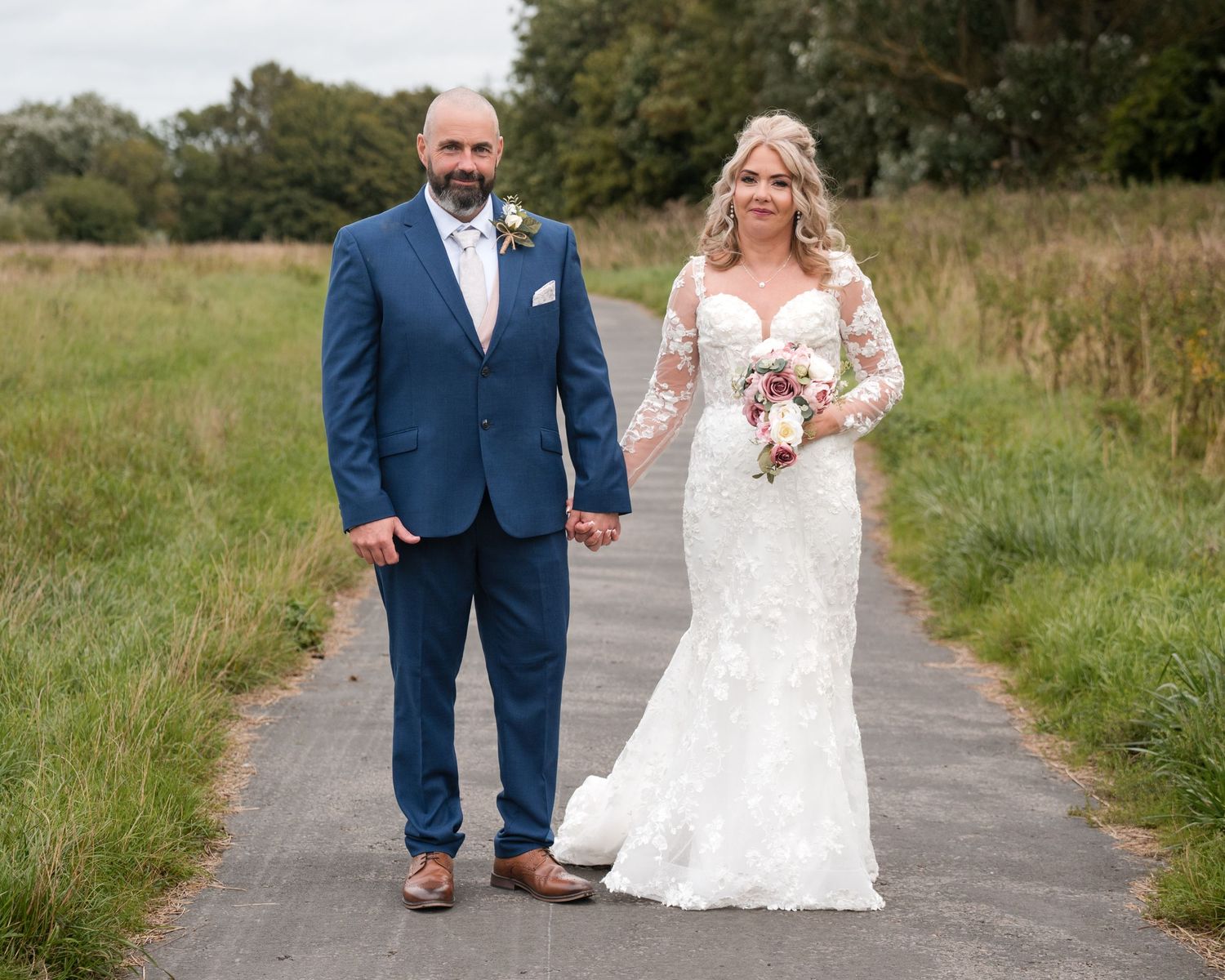 Bride in lace gown and groom in blue suit hold hands on a path surrounded by green fields on their wedding day.