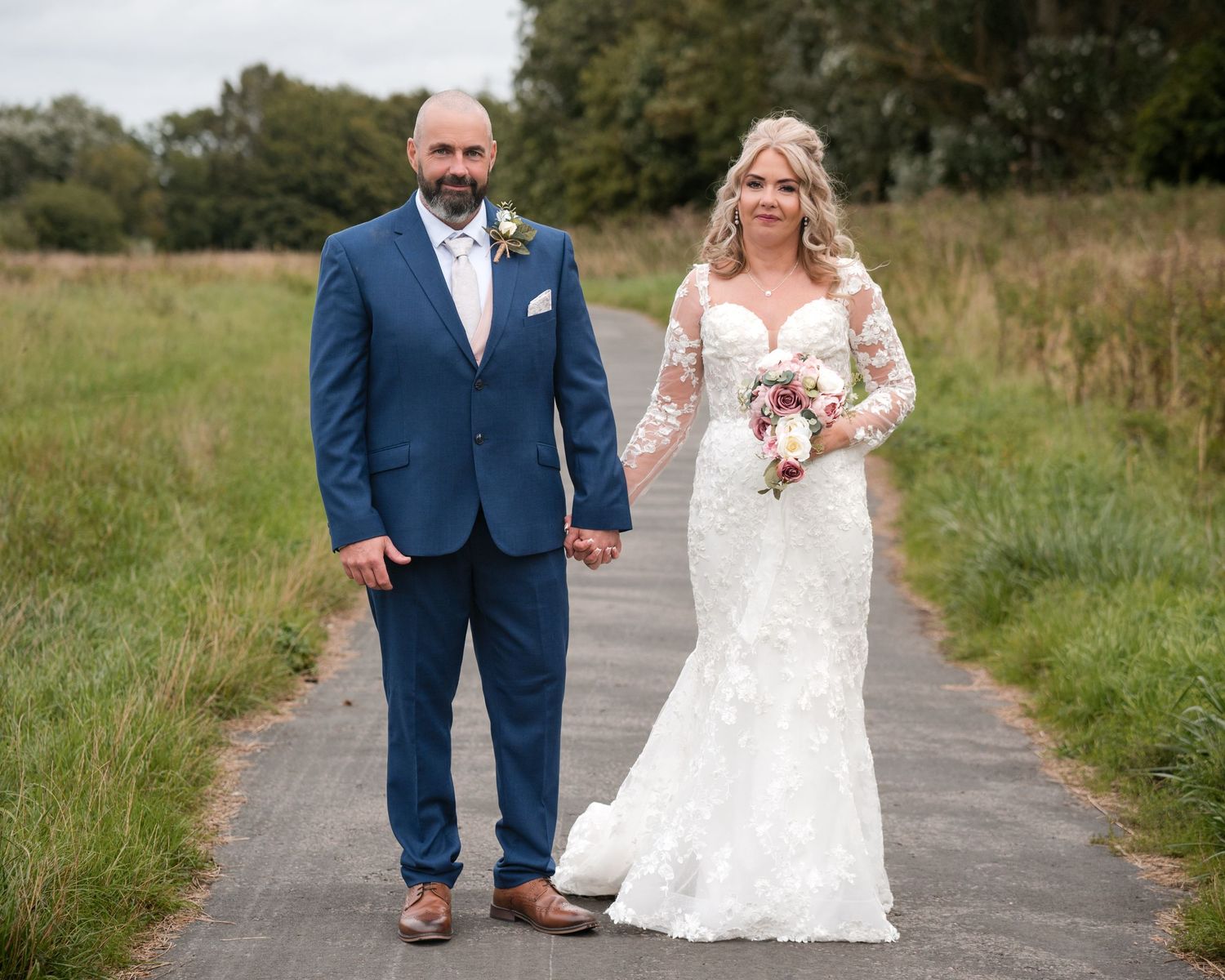 Bride in lace gown and groom in blue suit hold hands on a path surrounded by green fields on their wedding day.
