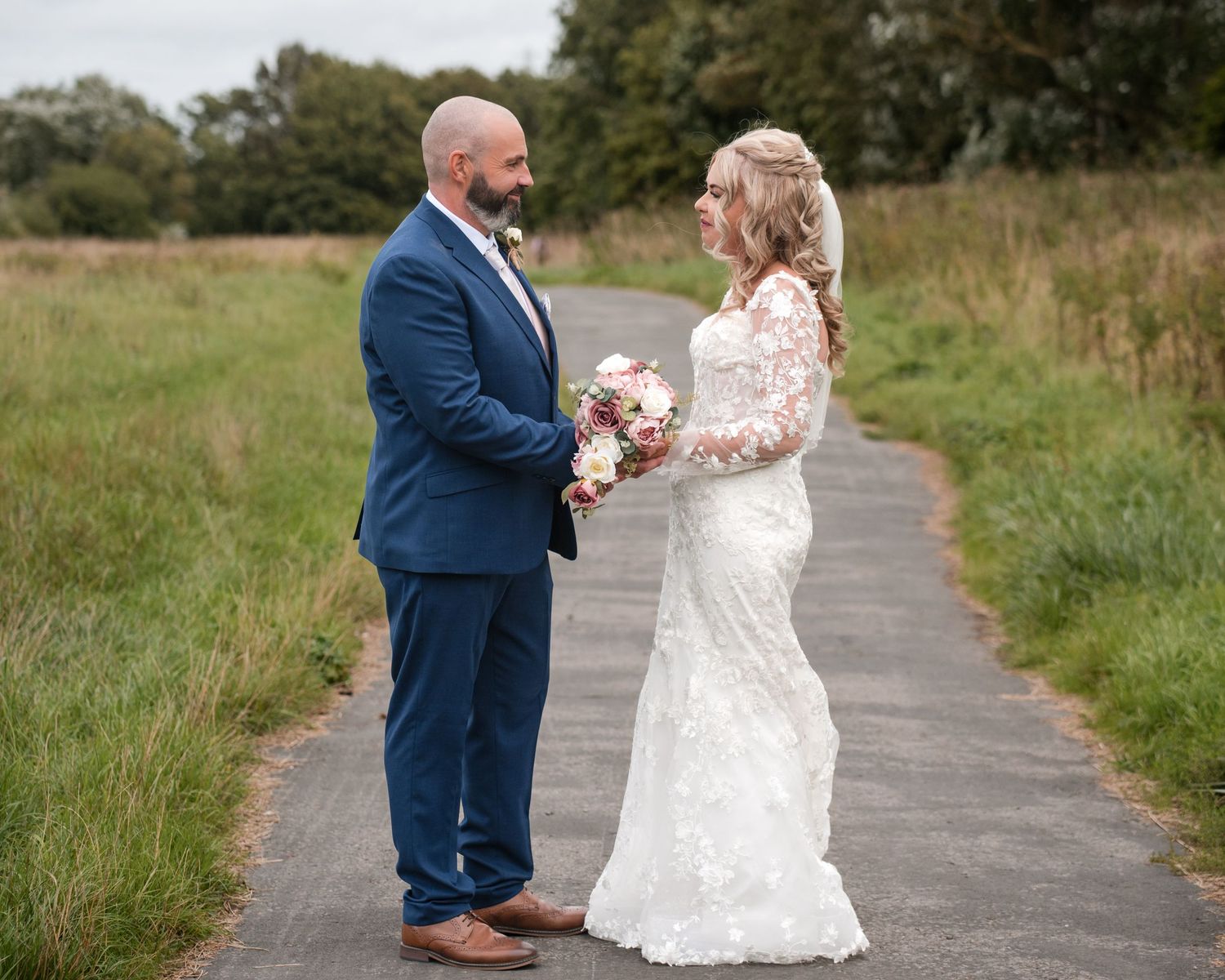 Bride and groom holding hands on a path, bride in lace gown with bouquet, groom in navy suit, surrounded by green fields.