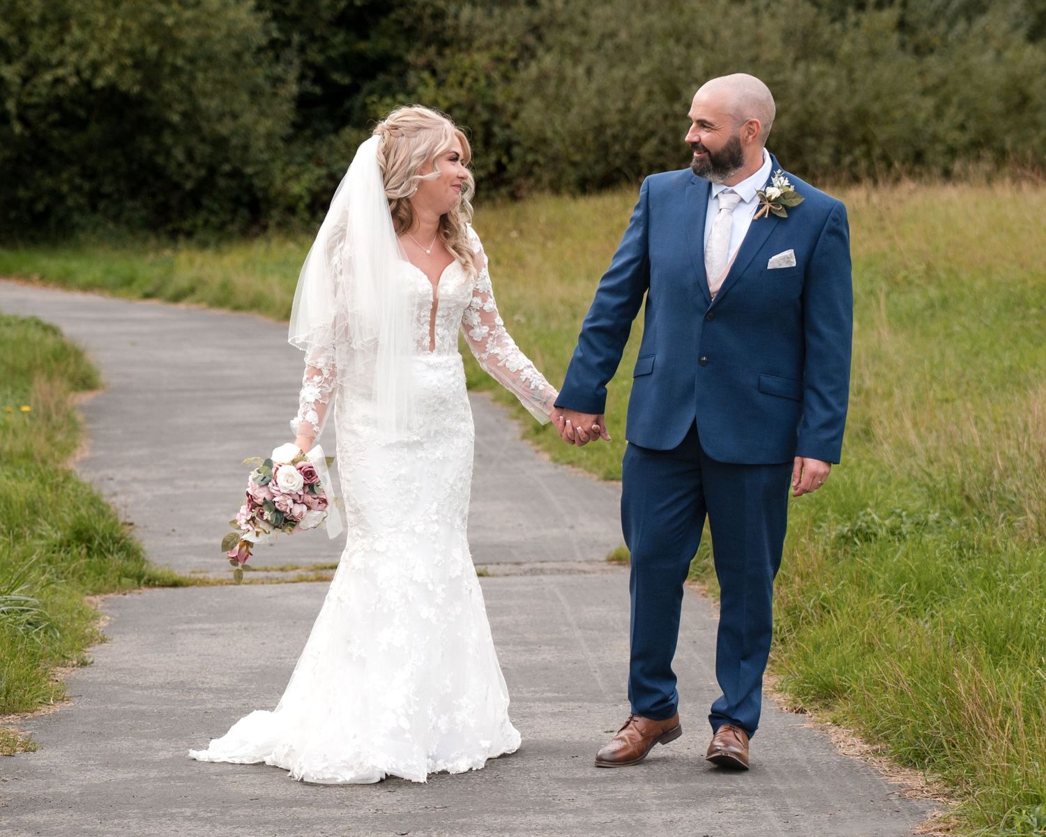 Bride in white lace gown and groom in navy suit hold hands on a path surrounded by green fields.