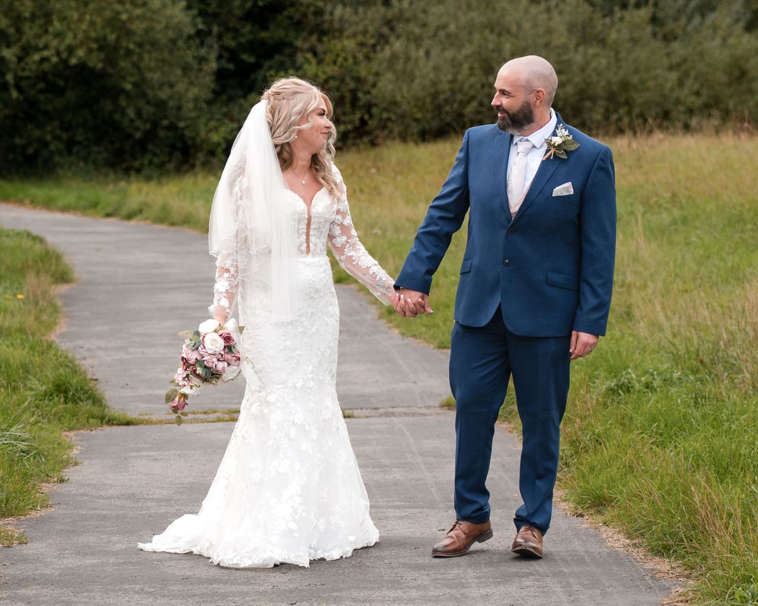 Bride in lace gown and groom in blue suit hold hands walking on path with green fields backdrop.