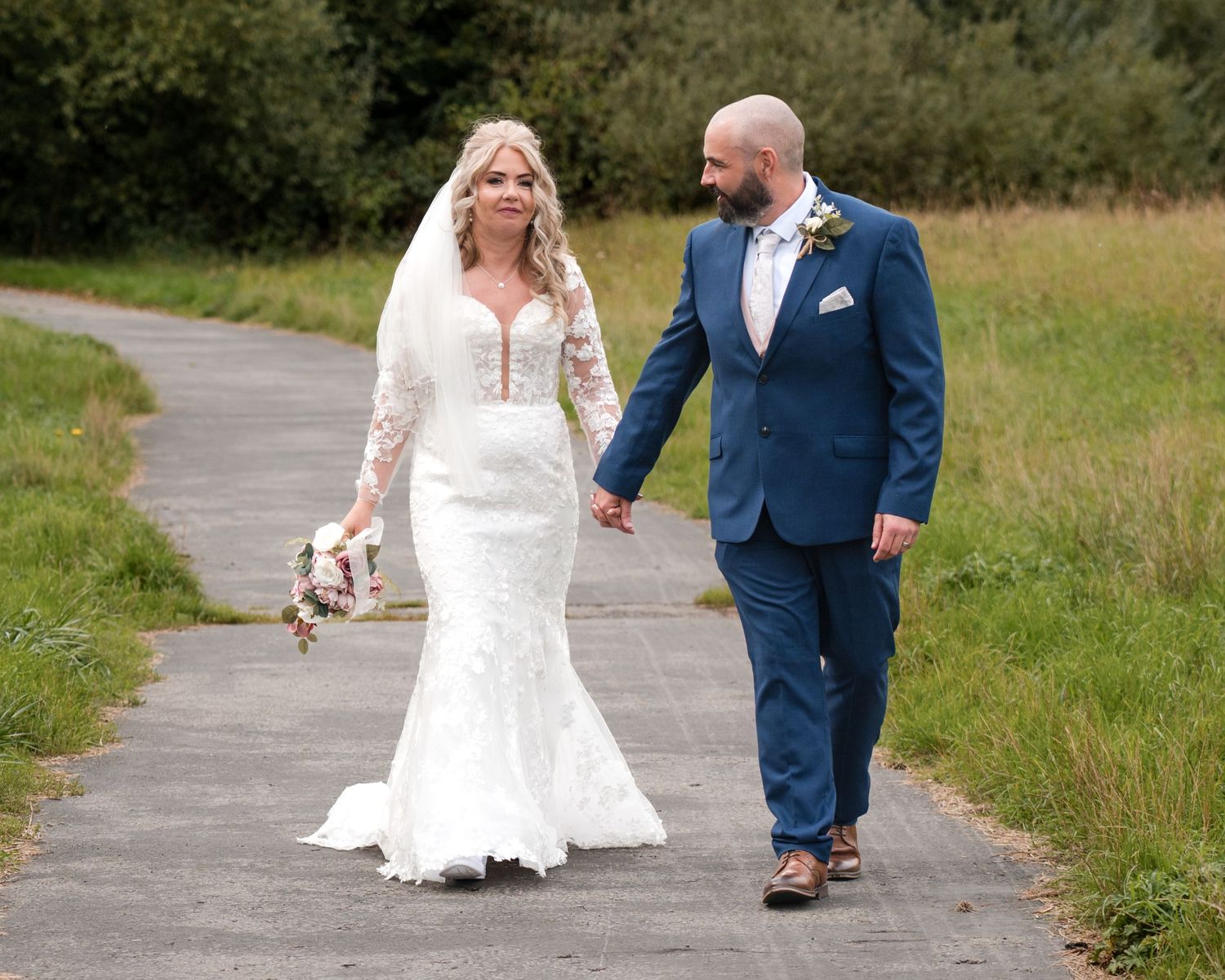 Bride in lace gown and groom in navy suit hold hands walking on path surrounded by green fields.