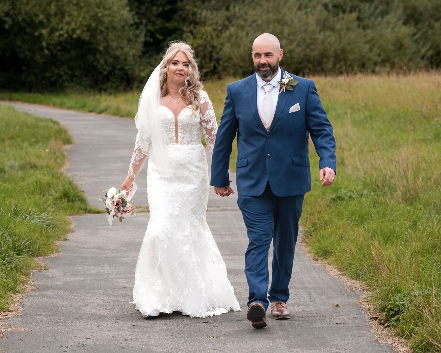 A bride and groom walk hand-in-hand on a path, she in a lace gown, he in a blue suit, on their wedding day.