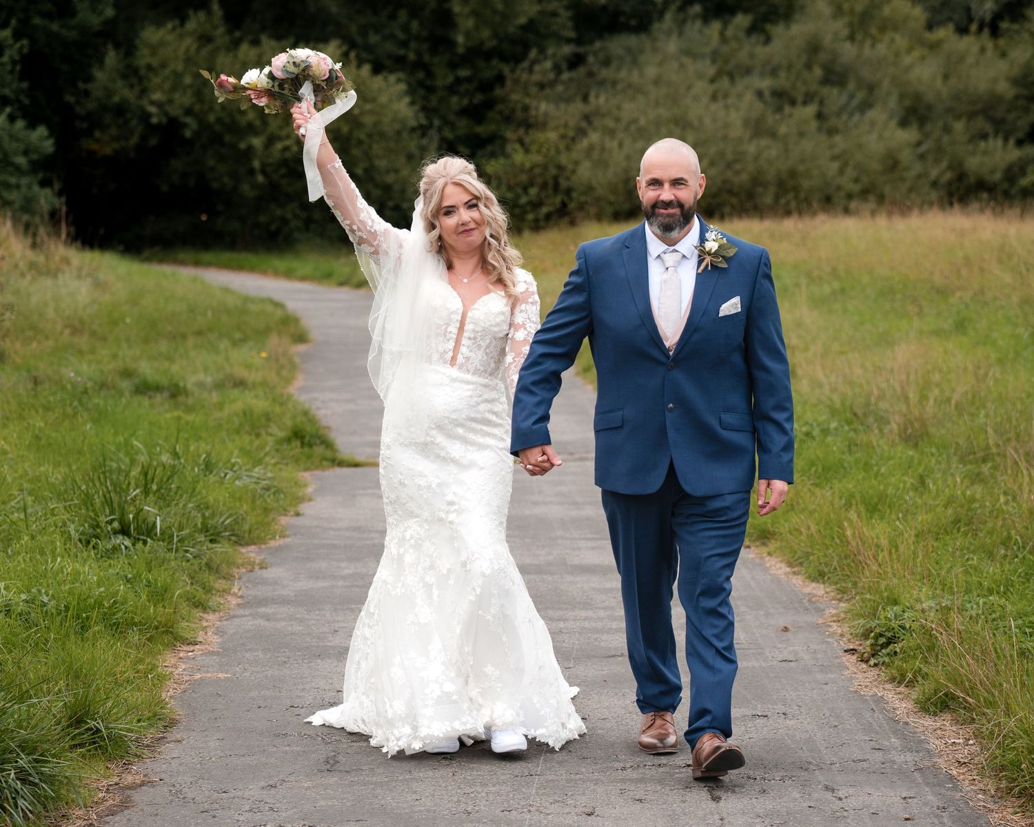 Bride in white lace gown raises bouquet while holding hands with groom in blue suit on a tree-lined path.