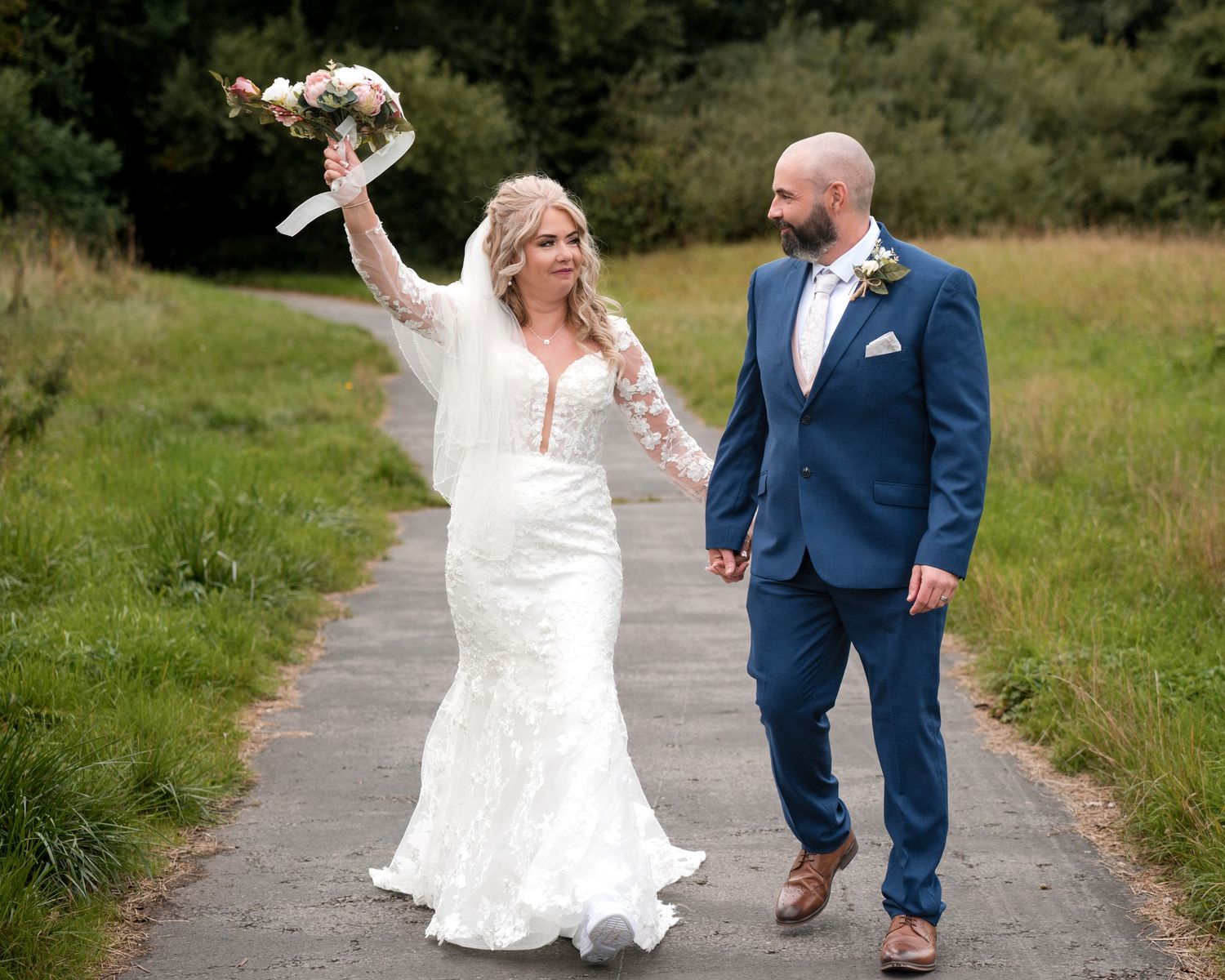 Bride in white lace gown raises bouquet while holding hands with groom in navy suit on a scenic outdoor path.