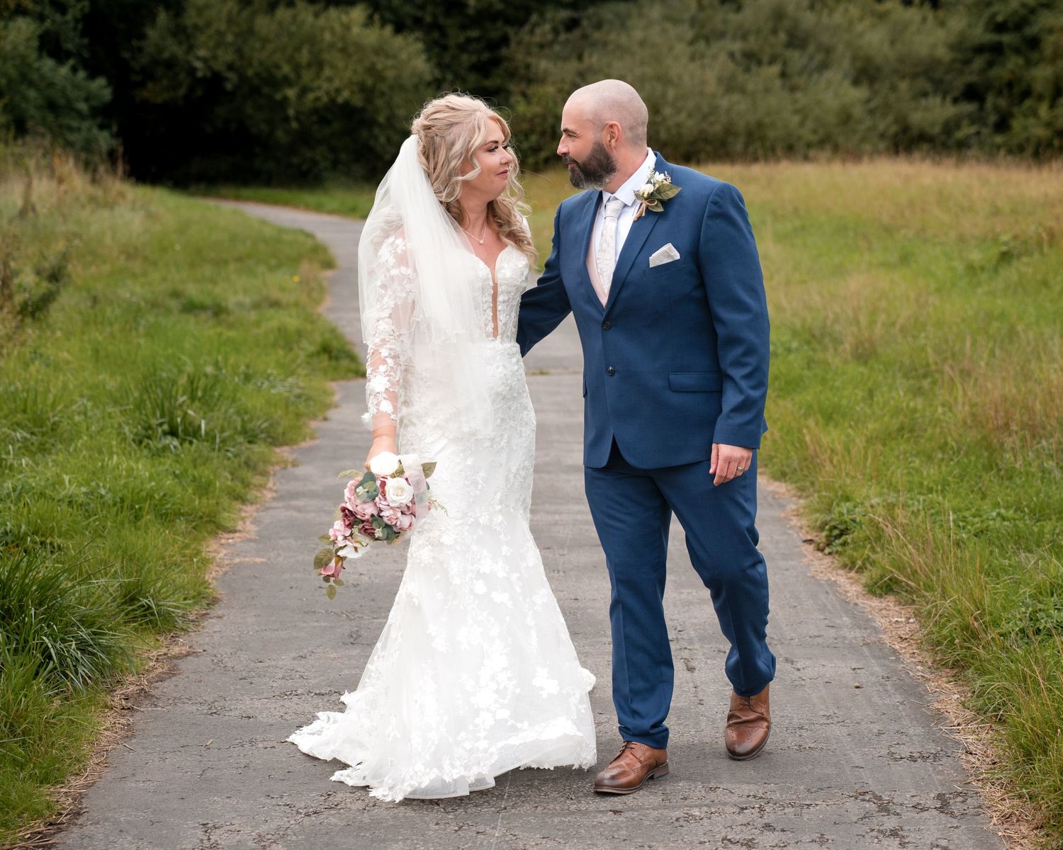 Bride in lace gown and groom in navy suit walk hand-in-hand on a path surrounded by lush greenery.