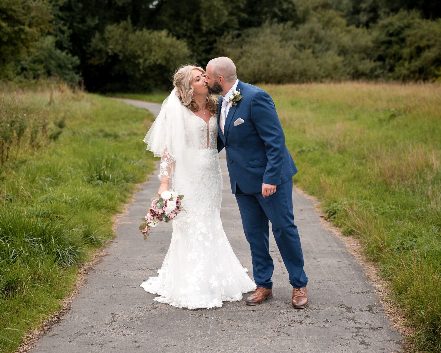Bride and groom kissing on a tree-lined path, bride in lace gown with bouquet, groom in navy suit.