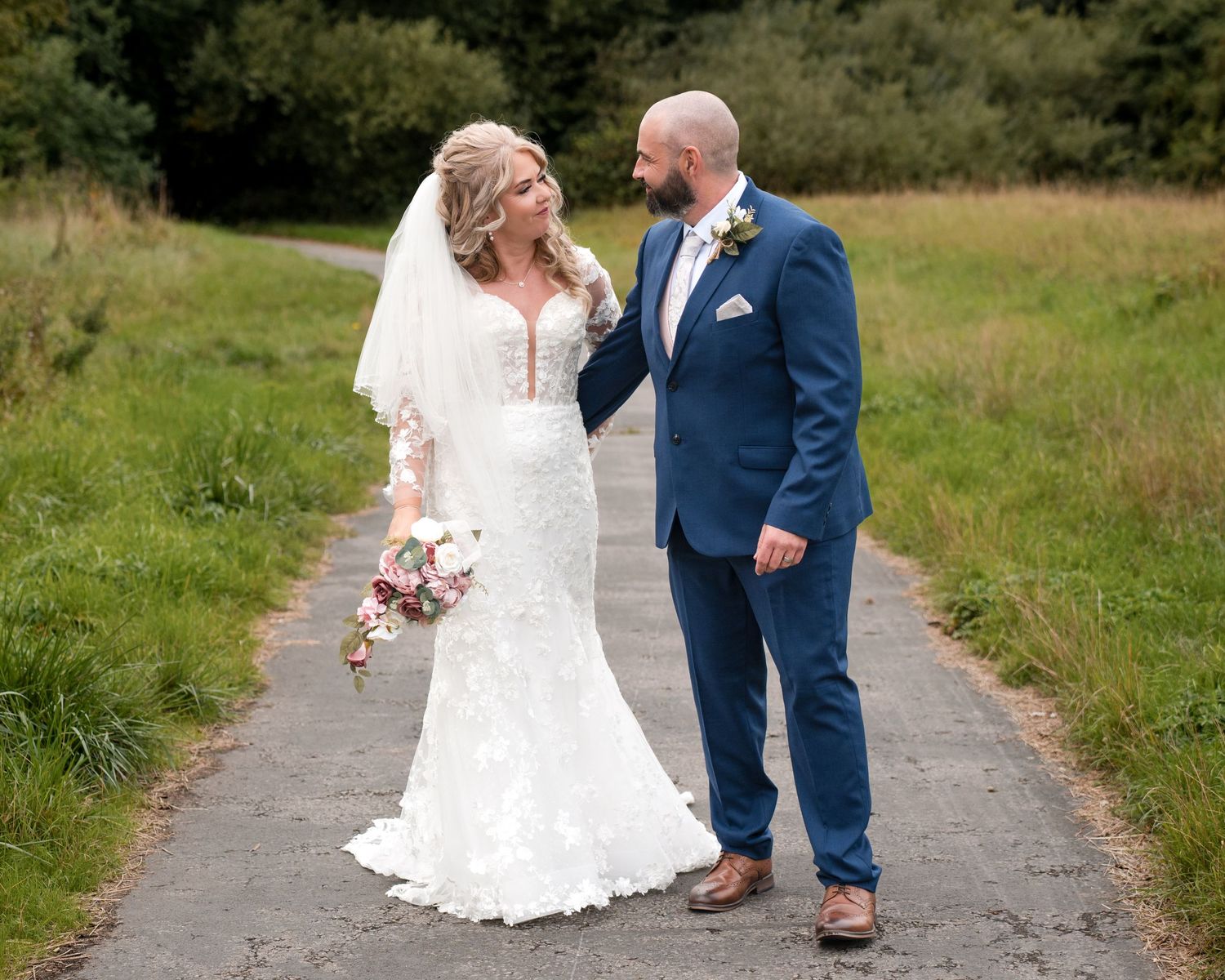 Bride in lace gown and groom in blue suit walk a garden path, exchanging glances on their wedding day.
