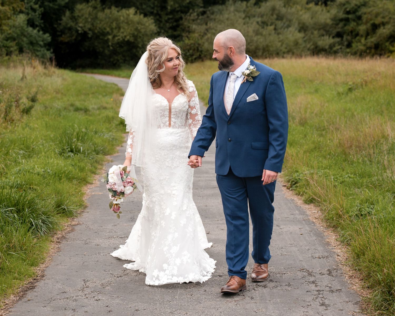 Bride in lace gown and groom in navy suit hold hands walking on a path surrounded by green fields.