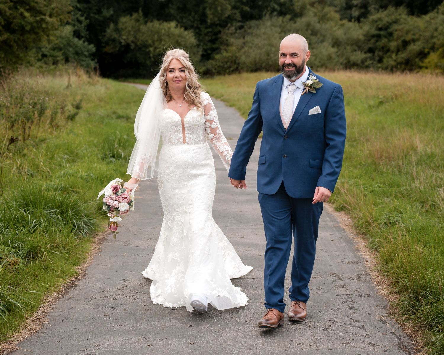 Bride and groom walking hand in hand on a path, she in a mermaid dress, he in a blue suit.