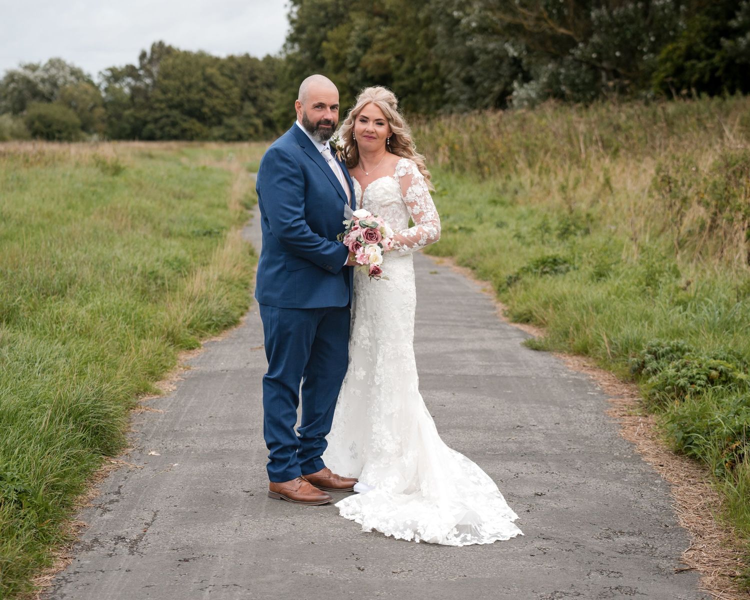 Bride in lace gown and groom in navy suit pose on a rustic path surrounded by green fields on their wedding day.