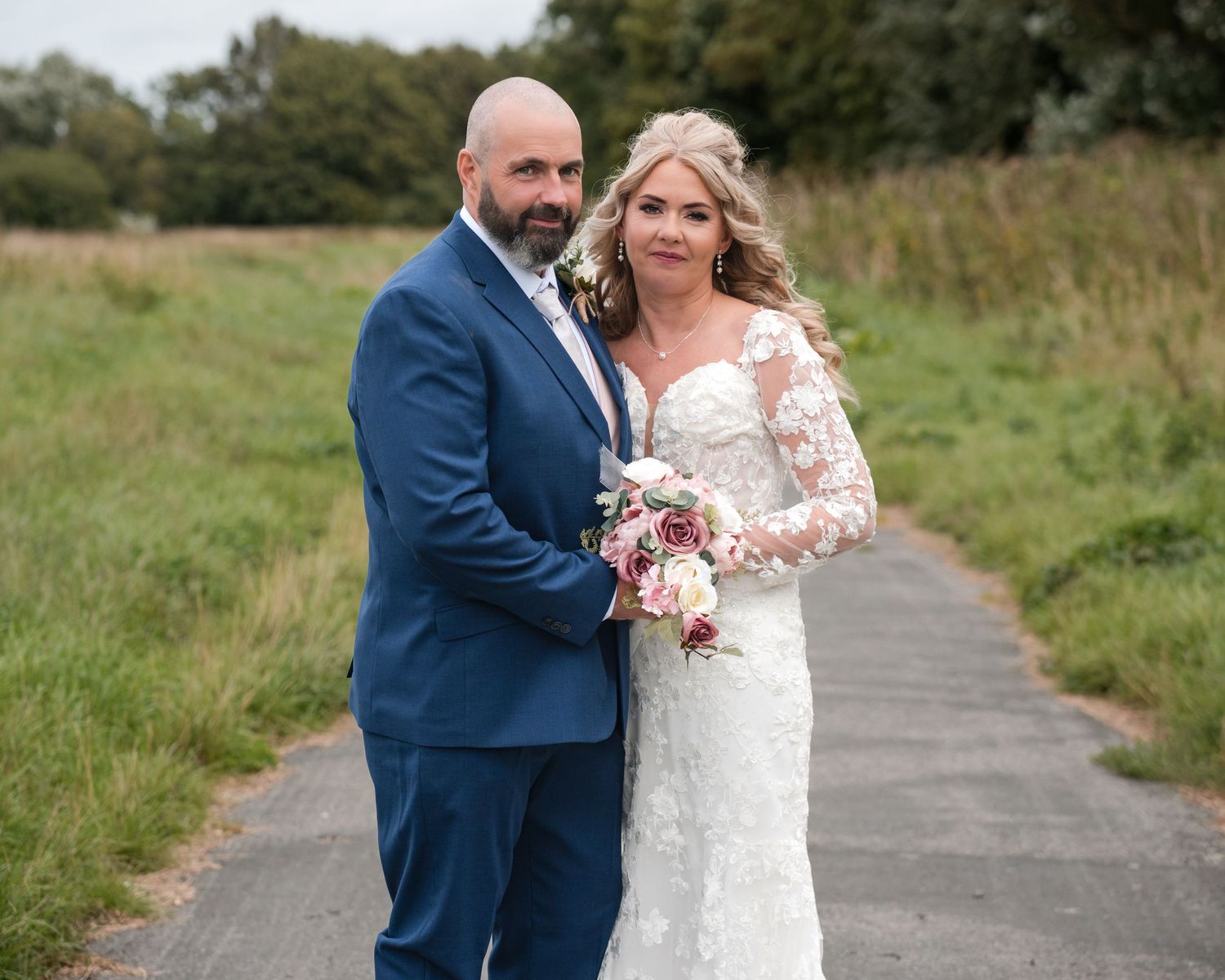Bride in lace gown and groom in navy suit pose on a path surrounded by green fields on their wedding day.