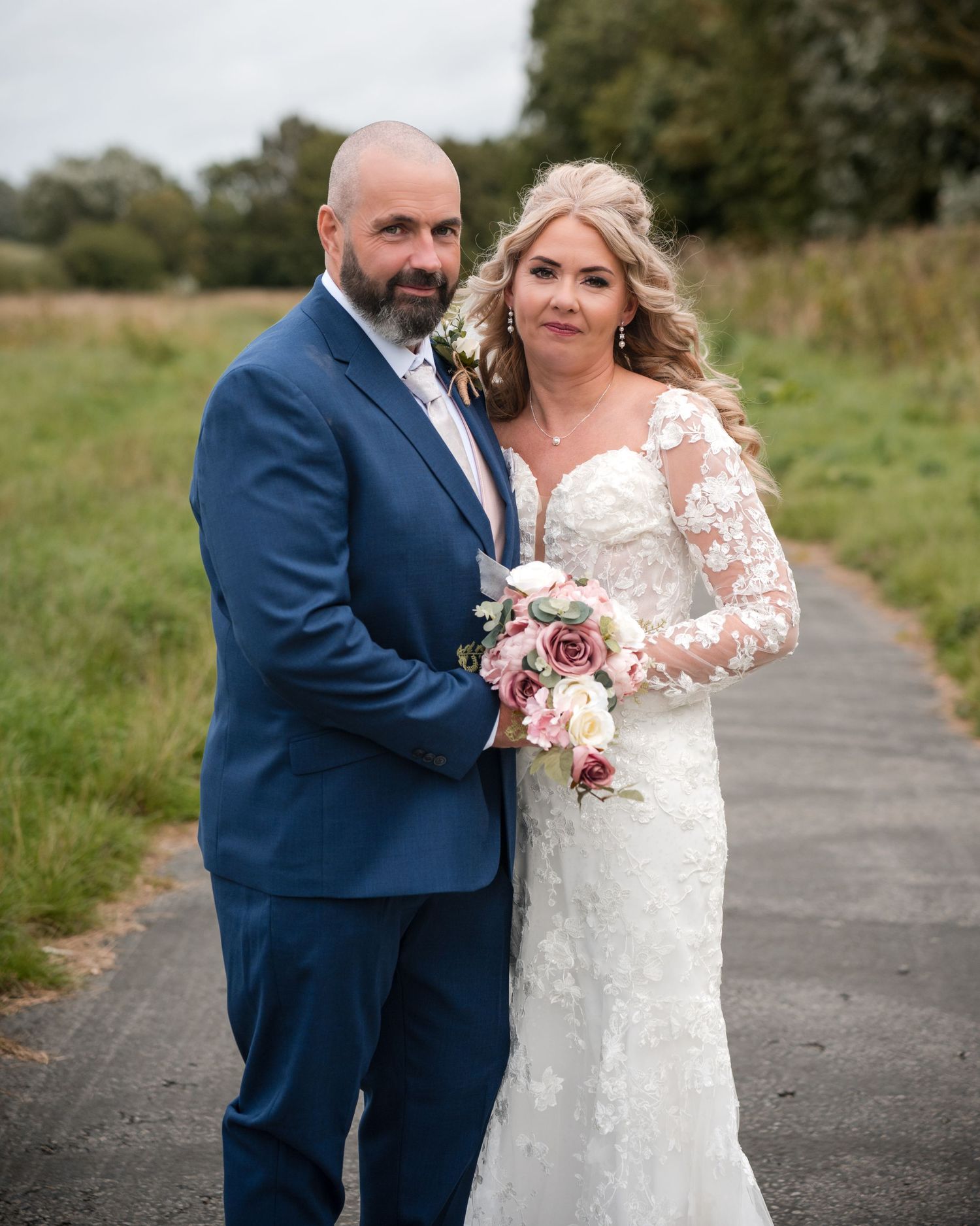 Bride in lace gown holding bouquet stands with groom in navy suit on a path surrounded by green fields.