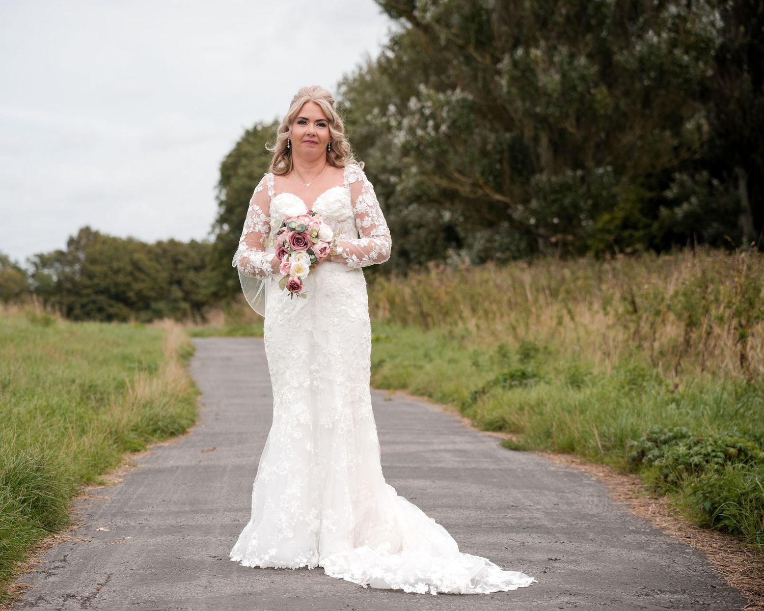 Bride in white lace gown with long sleeves holds bouquet on a paved path surrounded by green fields.