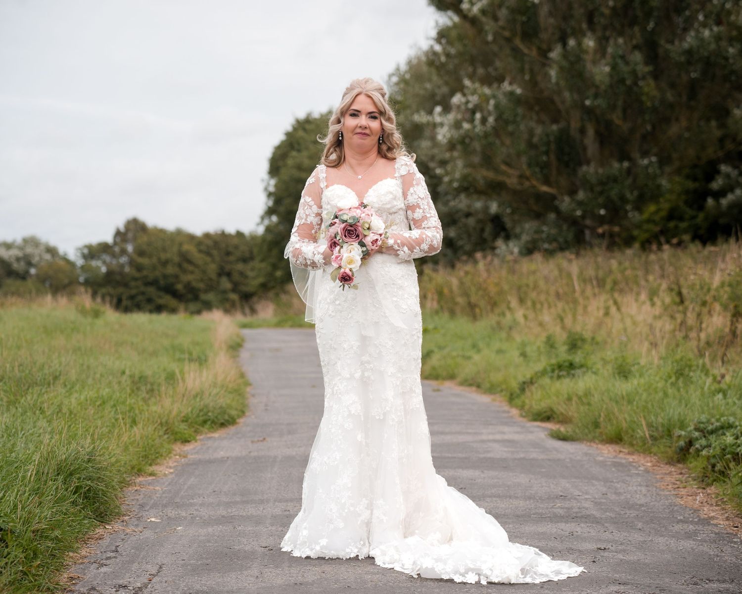 Bride in lace wedding dress holding bouquet standing on a path surrounded by green fields and trees.