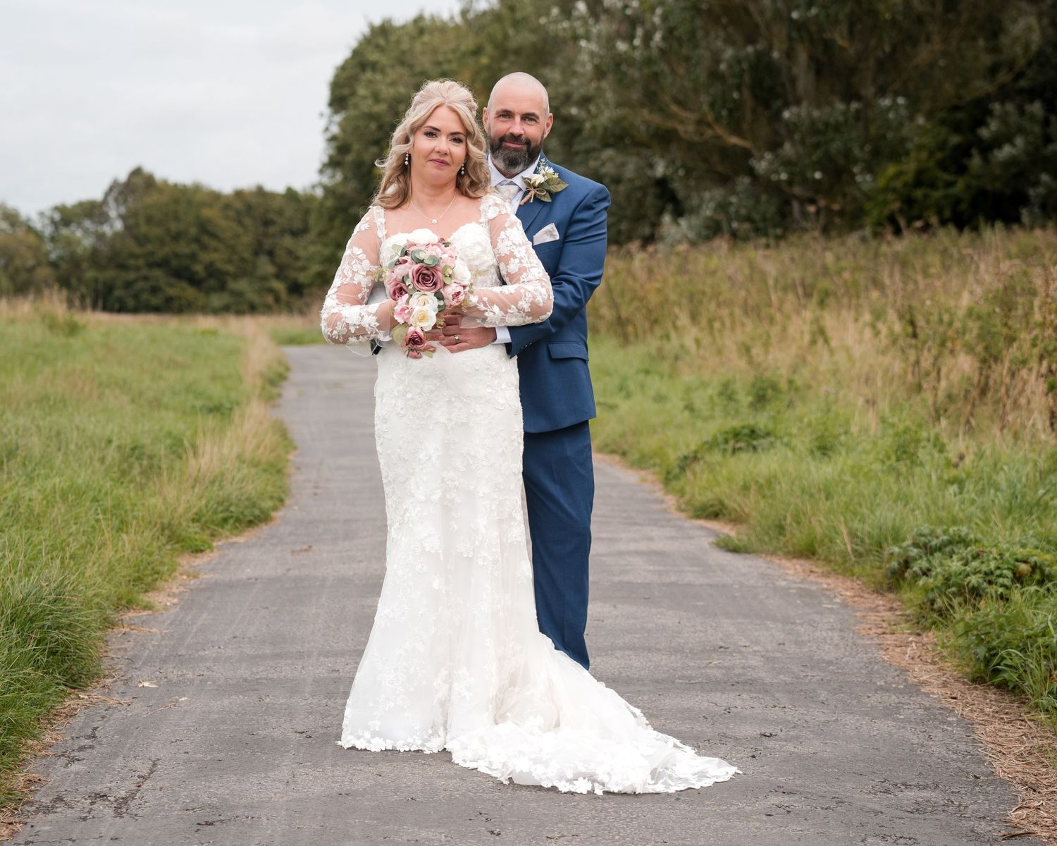 Bride in lace gown holding bouquet stands with groom in navy suit on a path surrounded by greenery.