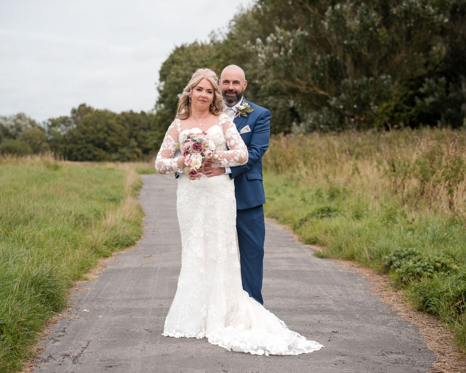 Bride in lace gown and groom in navy suit pose on a country path surrounded by green fields on their wedding day.