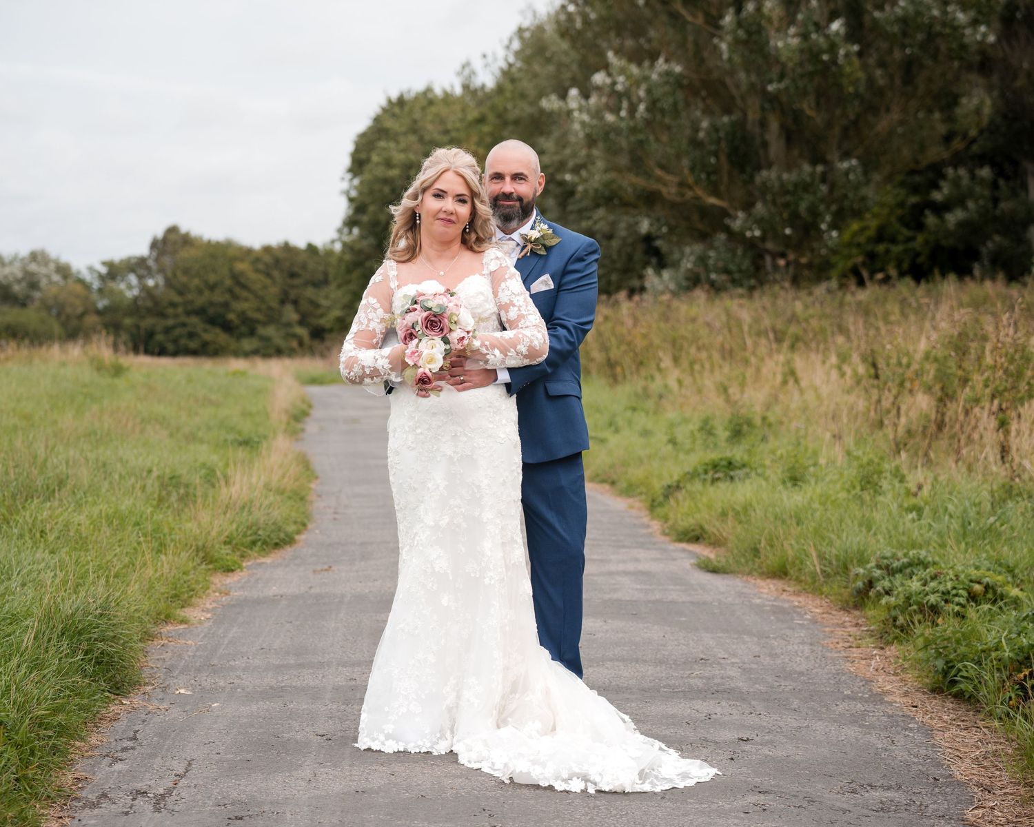 Bride in lace gown and groom in navy suit pose on a country path surrounded by green fields on their wedding day.
