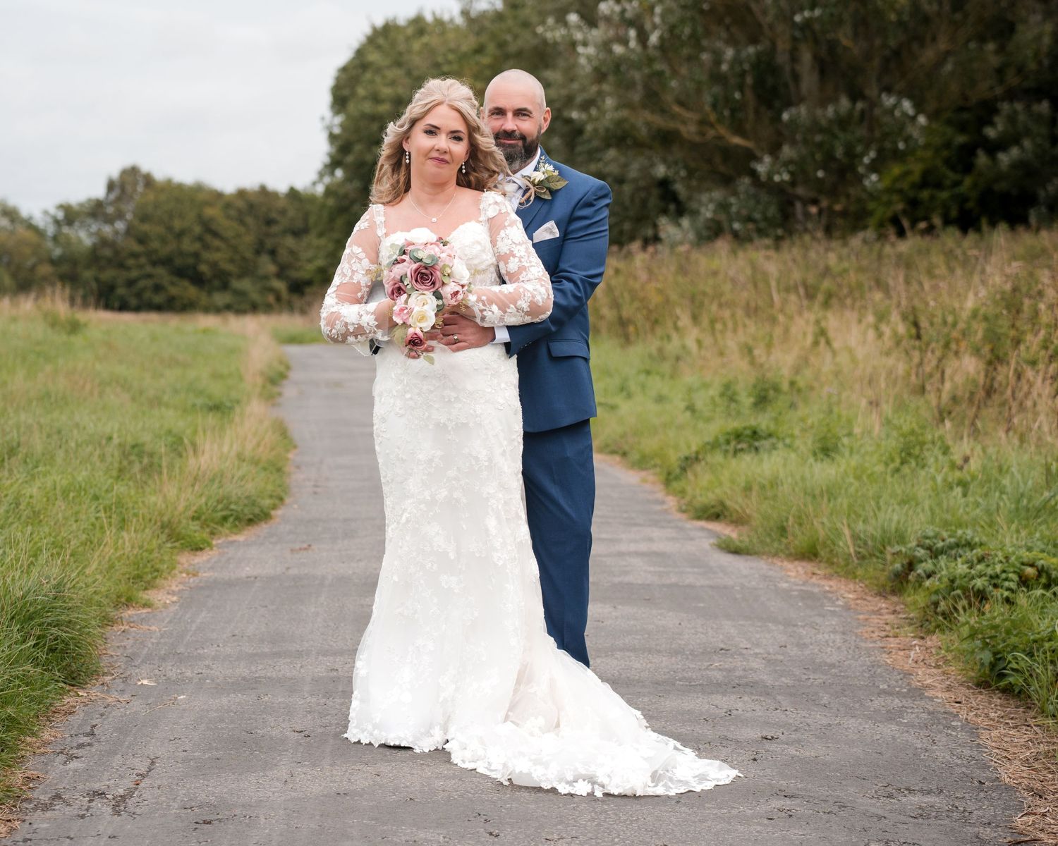 Bride in lace gown and groom in navy suit pose on a country path, surrounded by green fields on their wedding day.