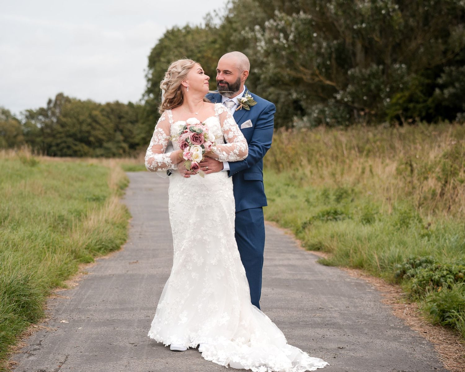 Bride in lace gown and groom in blue suit pose on a path surrounded by greenery on their wedding day.