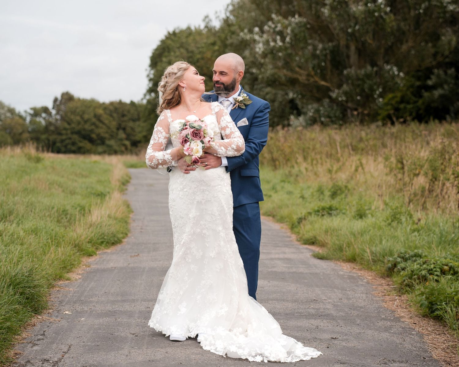Bride in lace gown and groom in navy suit pose on a path surrounded by green fields on their wedding day.