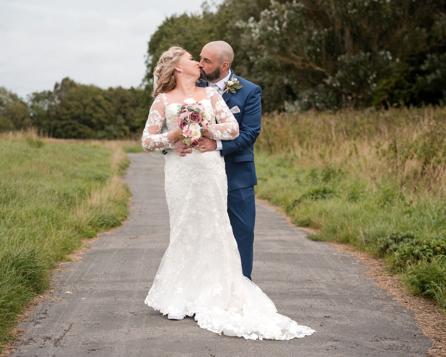 Bride in lace gown and groom in navy suit share a romantic moment on a path surrounded by greenery.