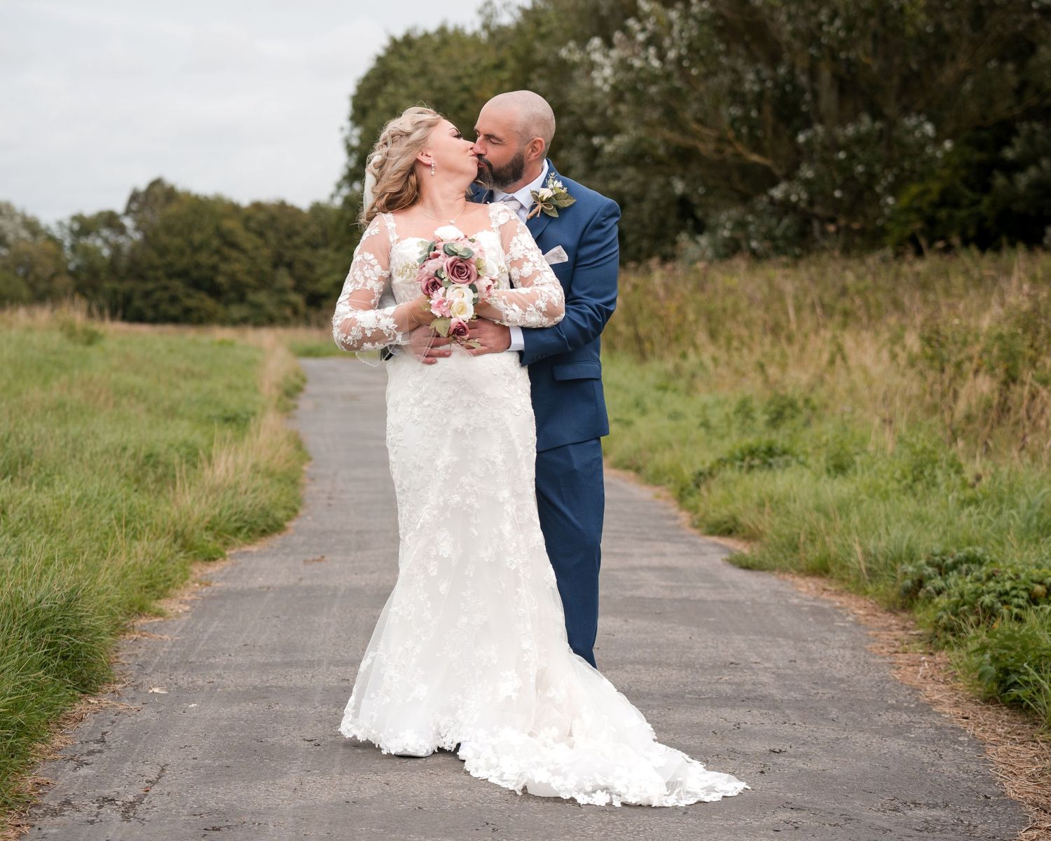 Bride in lace gown and groom in navy suit share a kiss on a country path surrounded by green fields.