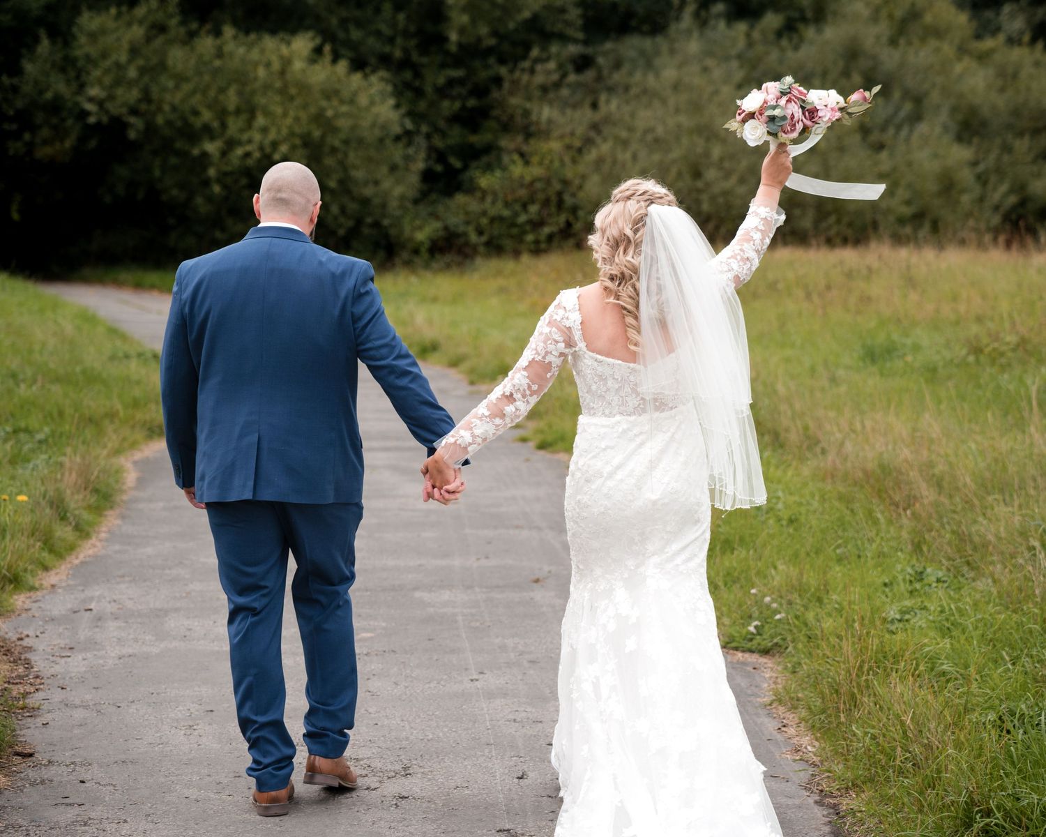 A bride and groom walk hand-in-hand down a path, bride holding a bouquet aloft, wearing a lace gown and veil.