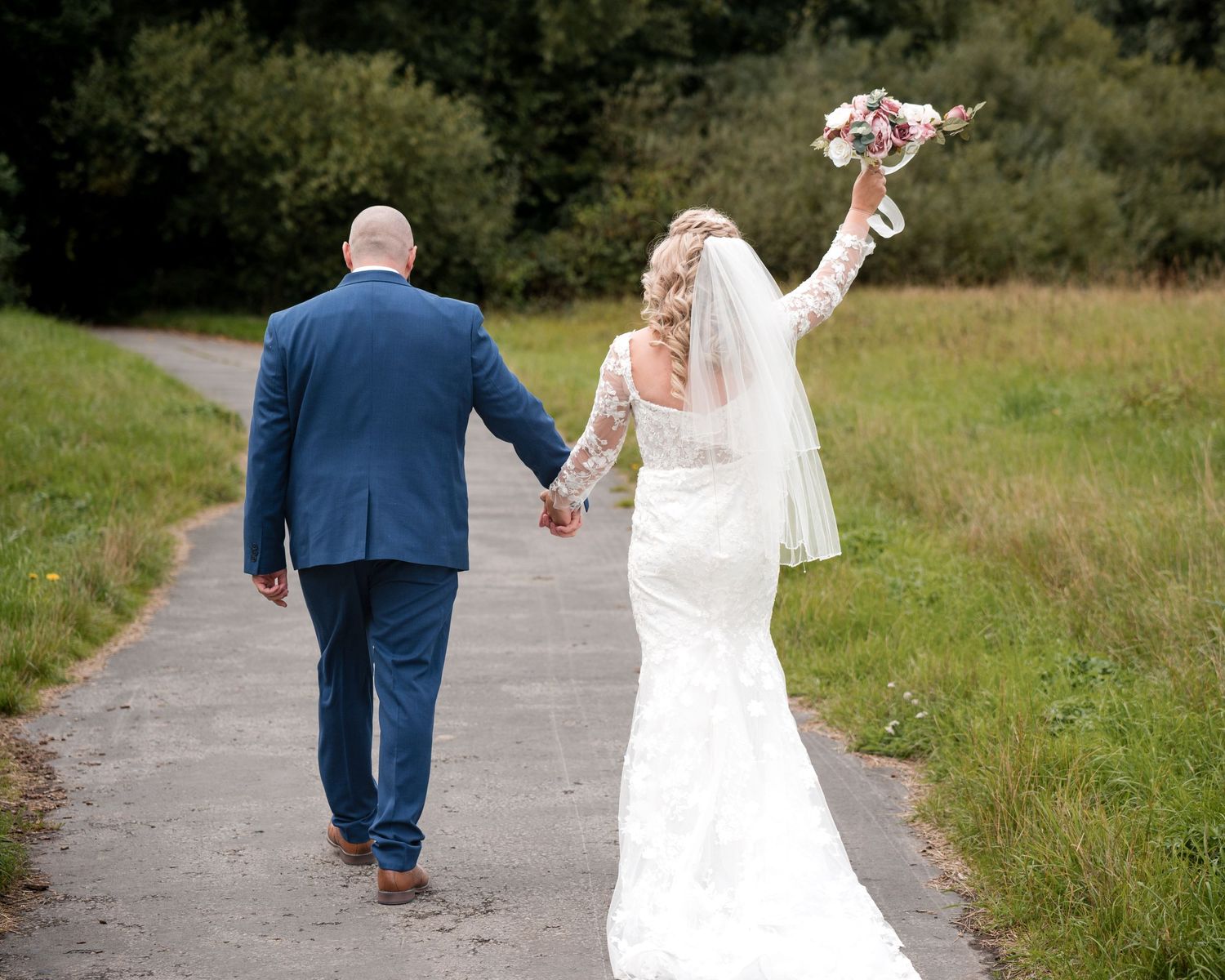 Bride and groom walking hand-in-hand down a path, bride raising bouquet, wearing lace dress and veil.