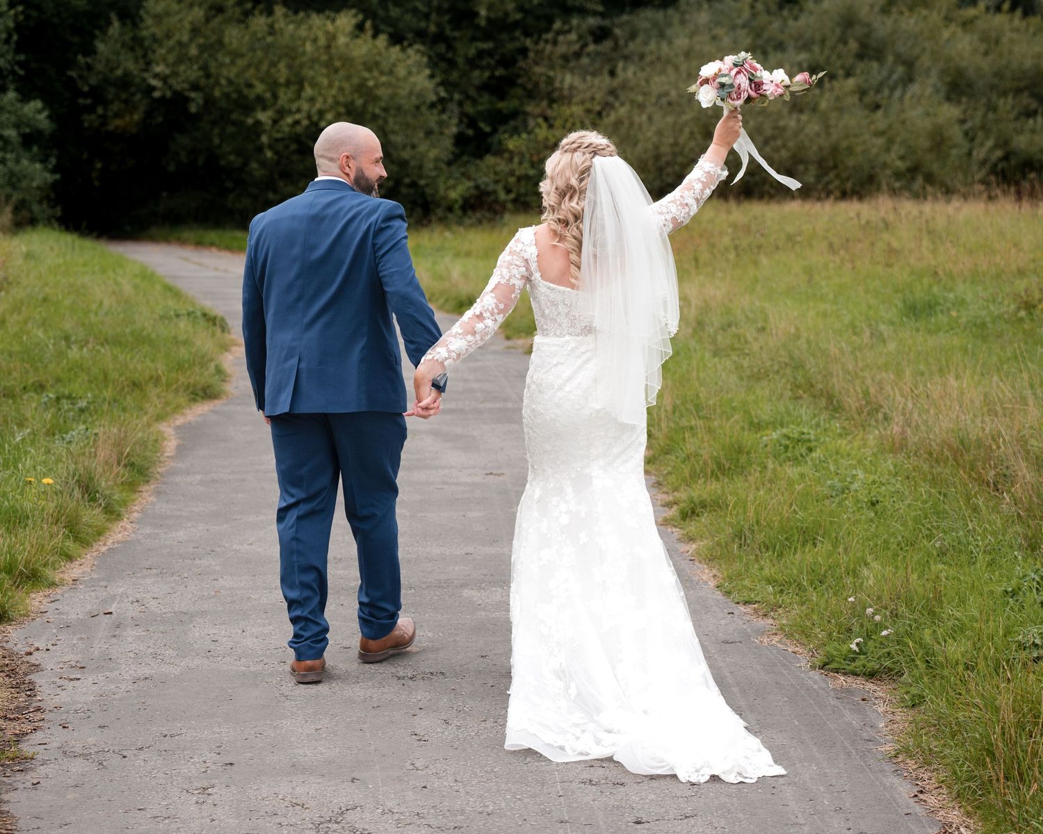 Bride in lace gown raises bouquet while holding hands with groom in blue suit walking down a path.