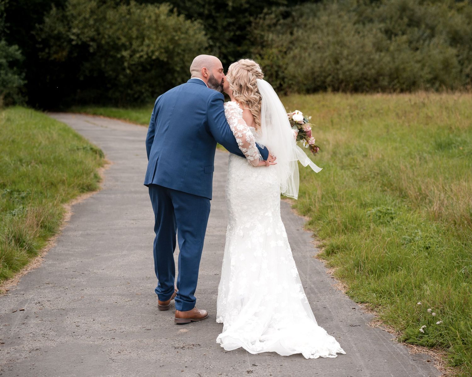 Bride and groom walking hand in hand down a path, woman in lace wedding dress holding bouquet, man in blue suit.