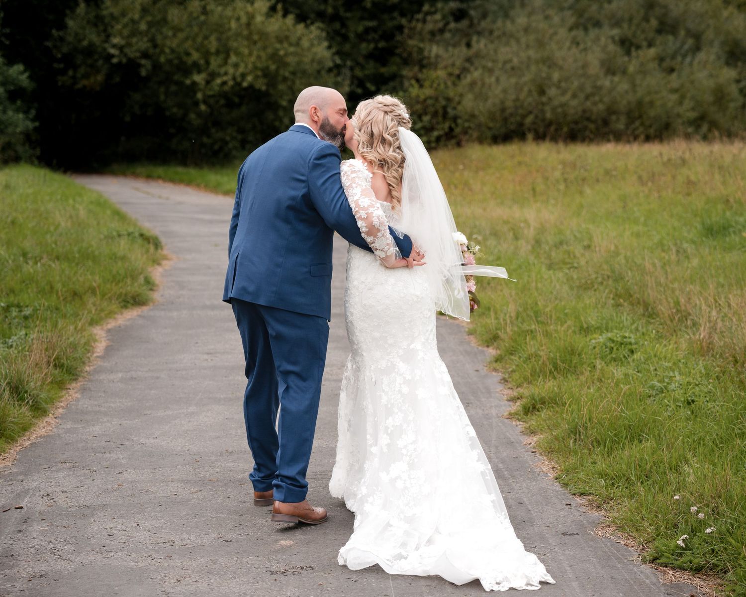Bride and groom walking hand in hand on a path, bride in lace gown with veil, groom in blue suit.