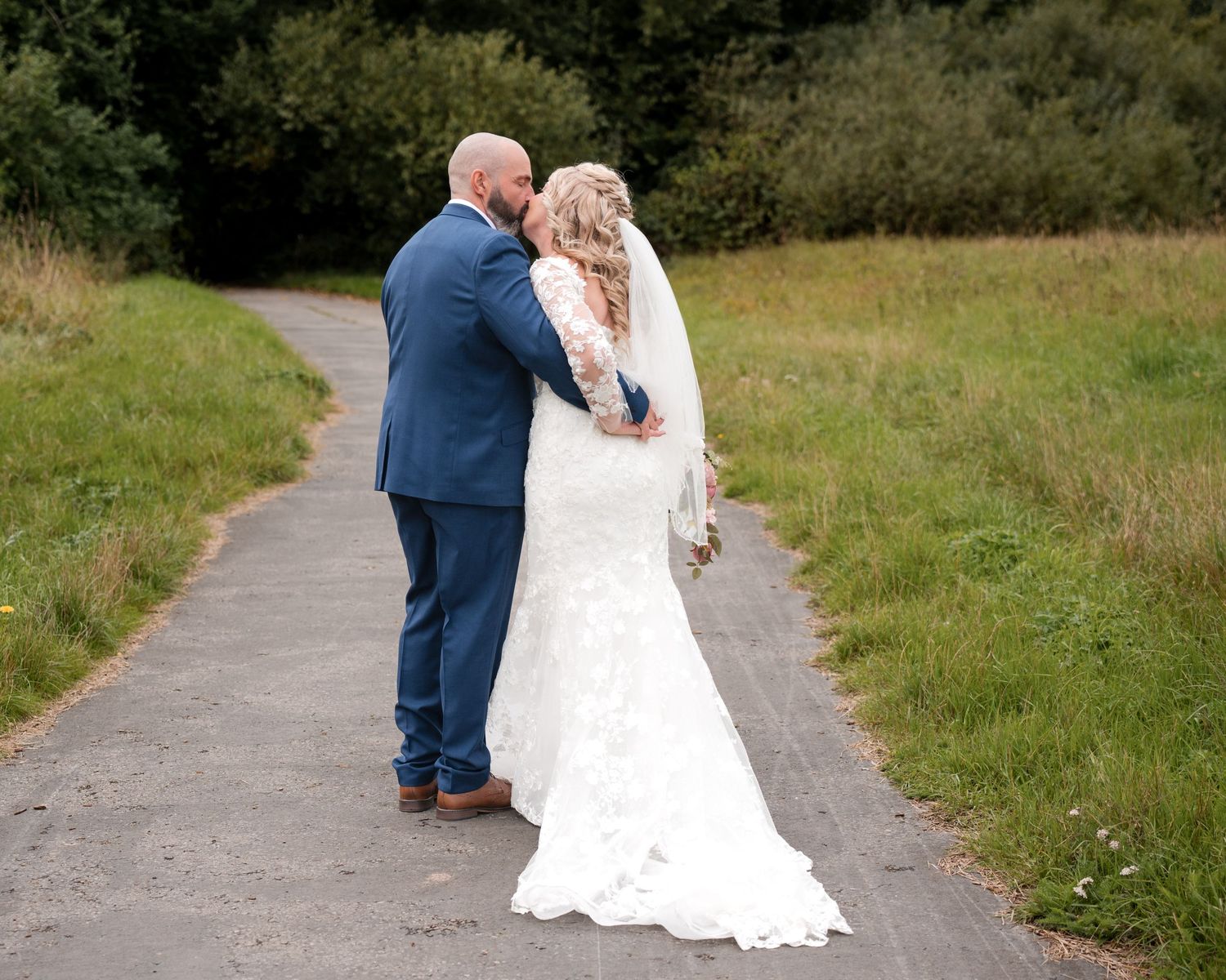 A bride and groom embrace on a path surrounded by greenery, the bride wearing a lace gown and veil.