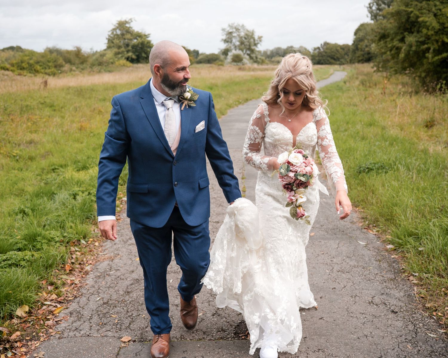 Bride and groom walking hand in hand on a country path, bride in lace gown holding bouquet, groom in blue suit.