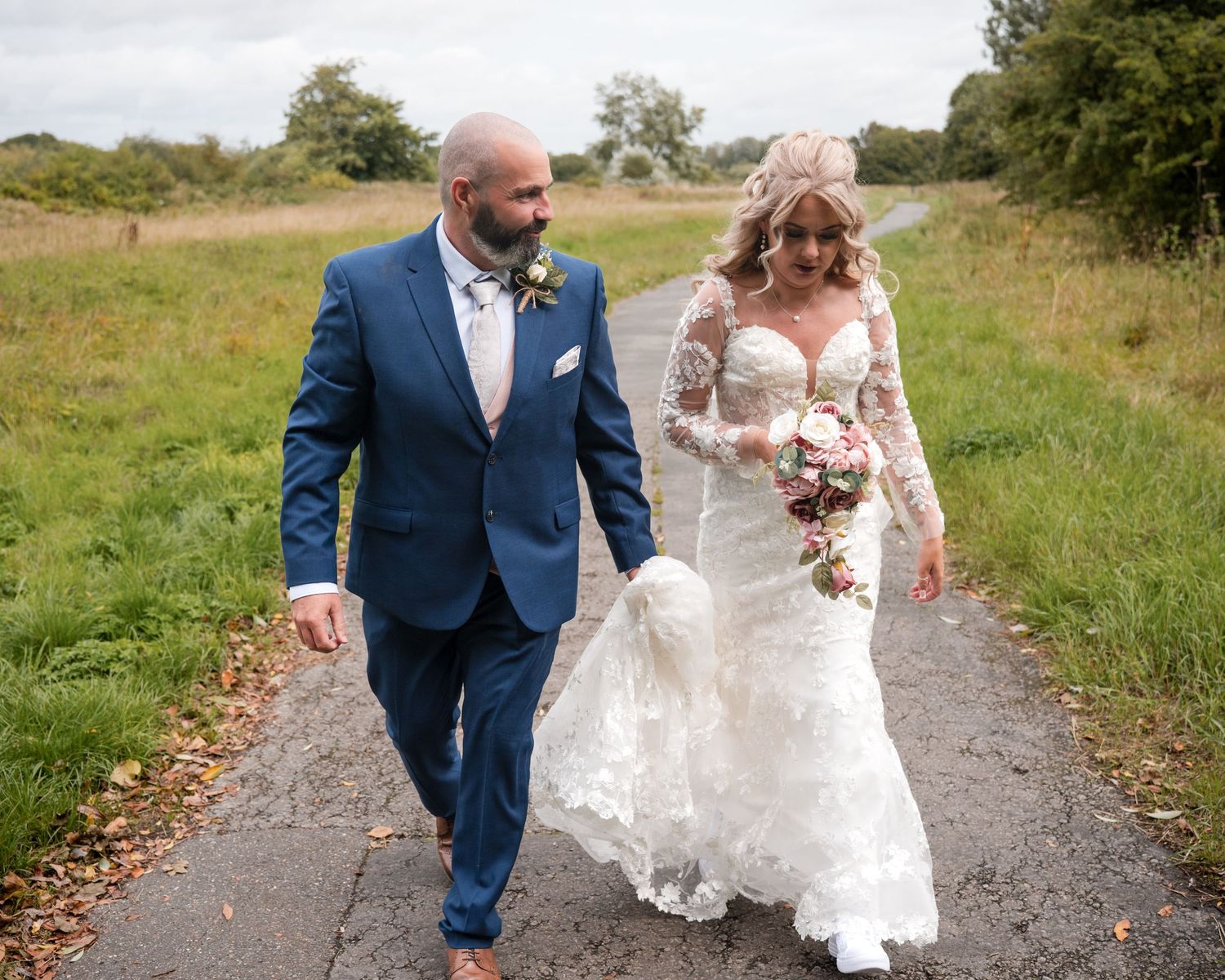 Bride and groom walking hand in hand on a country path, bride in lace gown, groom in blue suit.
