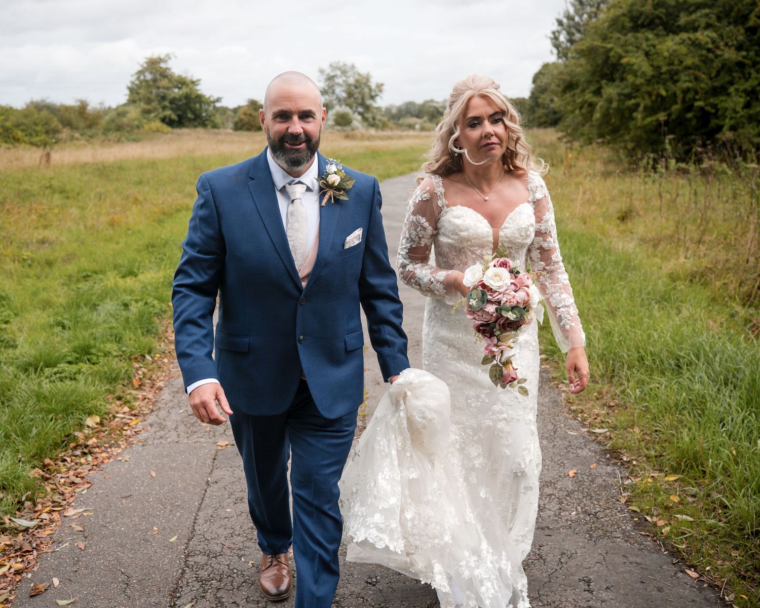 Bride in lace gown and groom in blue suit walk hand in hand along a path through green countryside fields.