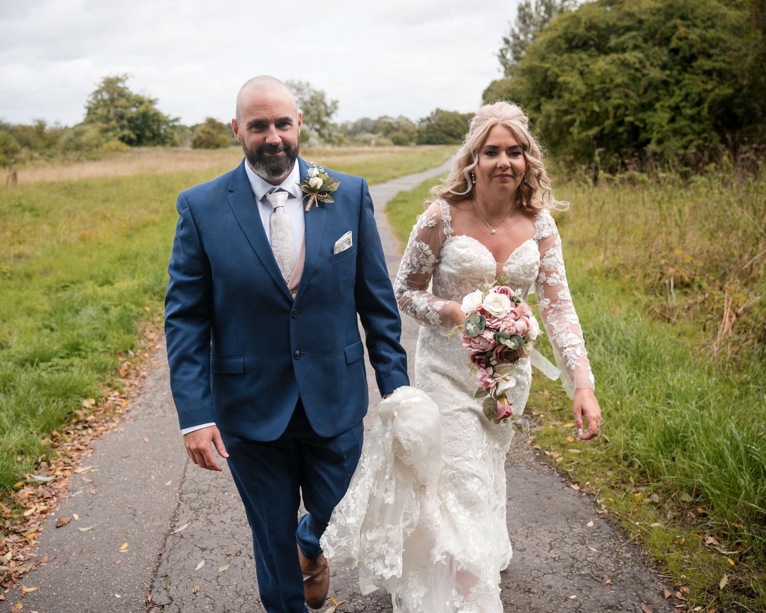 Bride in lace gown and groom in blue suit walk hand-in-hand on a path through green fields on their wedding day.