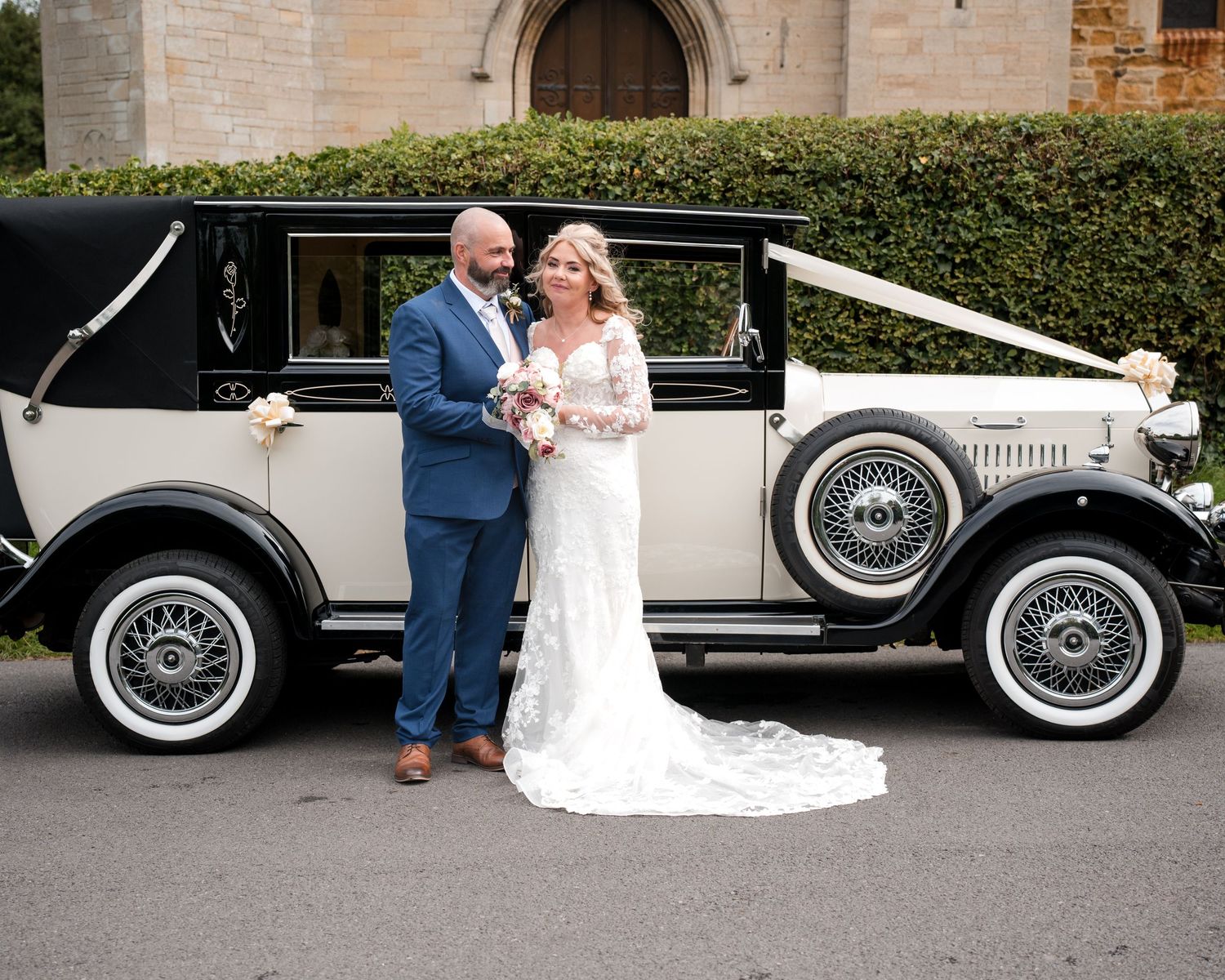 Bride and groom posing beside a vintage black and cream wedding car decorated with white ribbons outside a church.