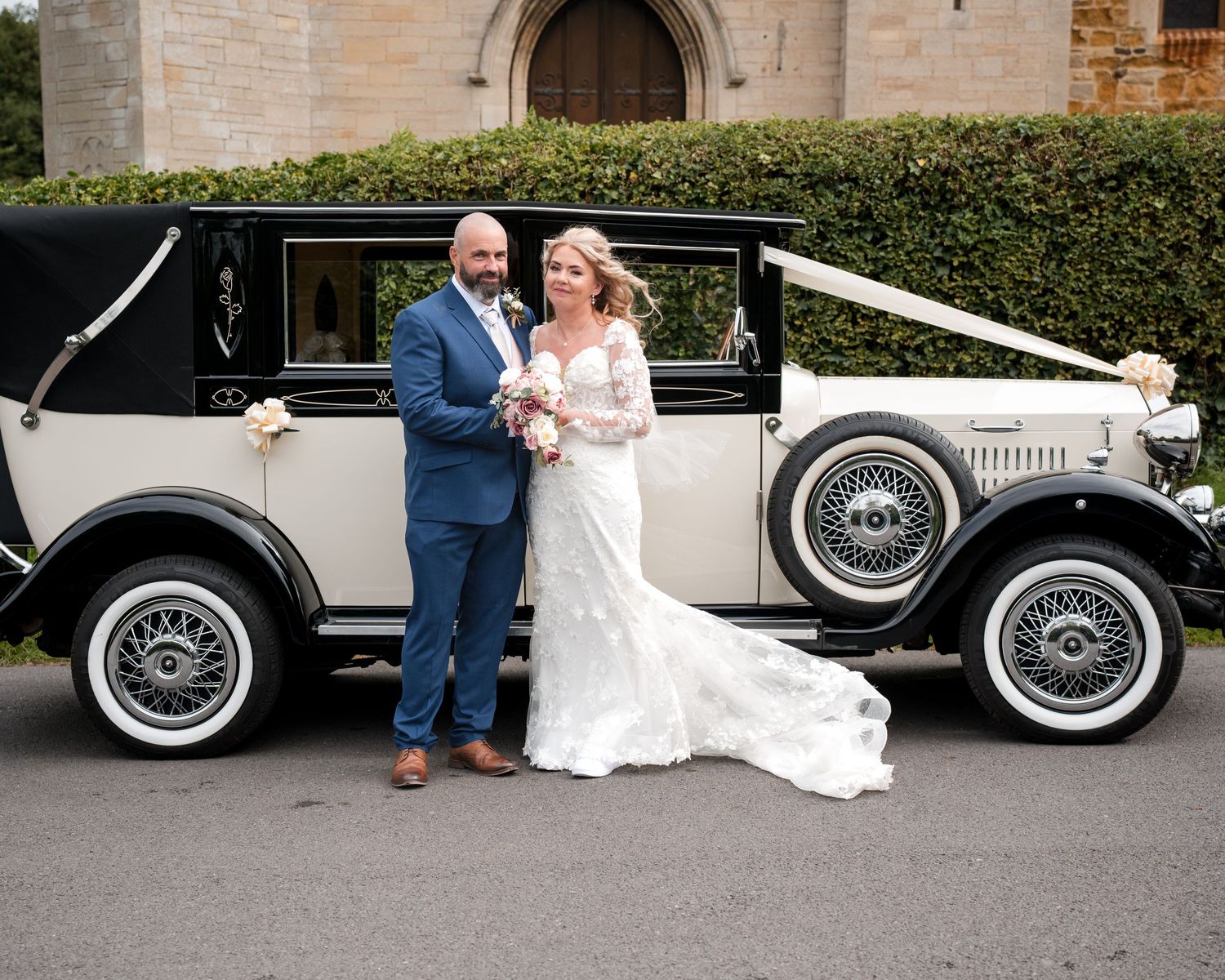 Bride and groom posing beside a vintage black and white wedding car decorated with white ribbons outside a church.
