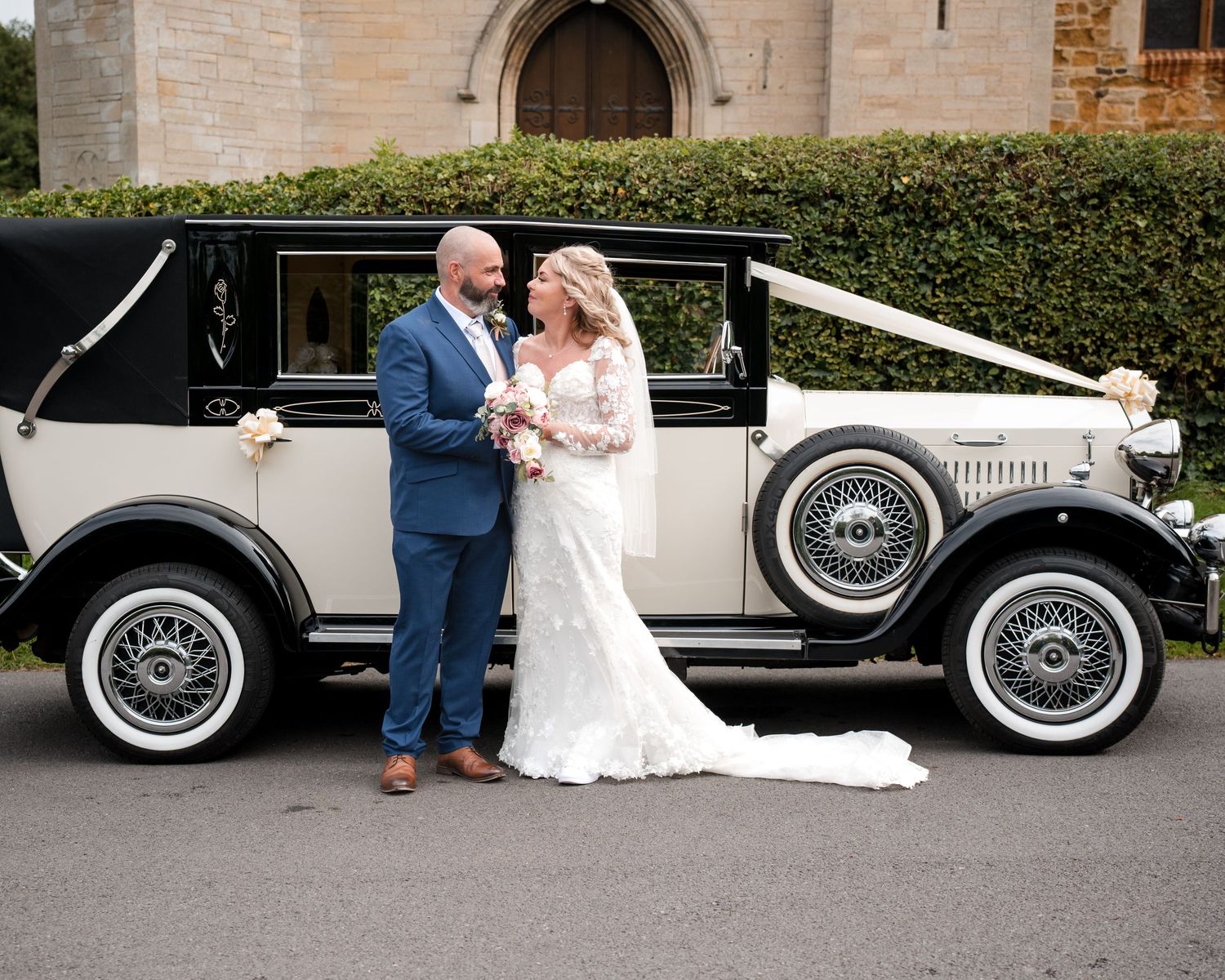 Bride and groom posing beside a vintage black and white wedding car decorated with ribbons outside a church.