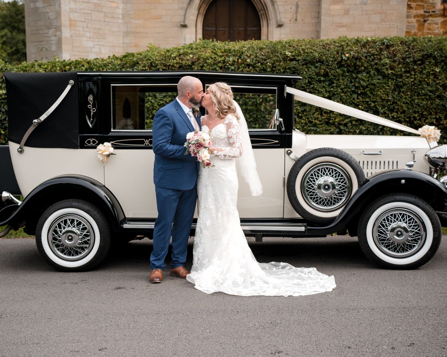 Bride and groom share a romantic moment beside a vintage black and cream wedding car adorned with white ribbons.