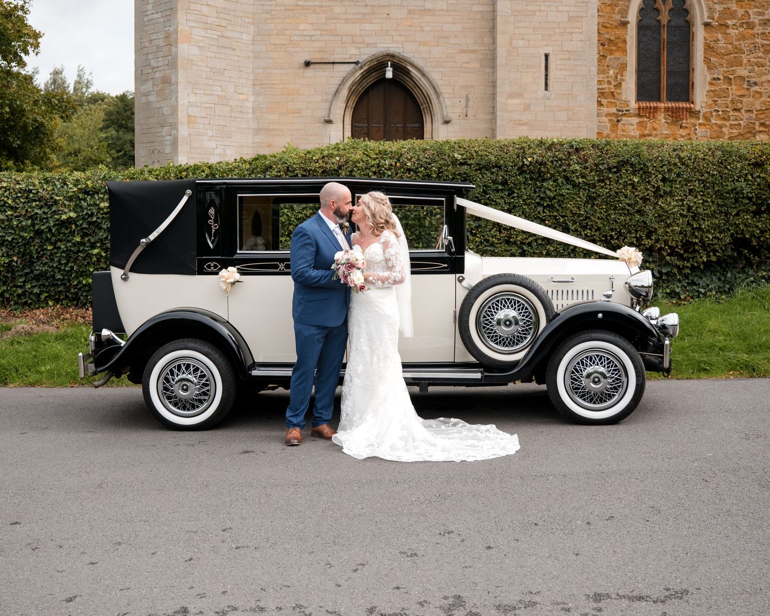 Bride and groom pose beside a vintage ivory wedding car outside a historic stone church.