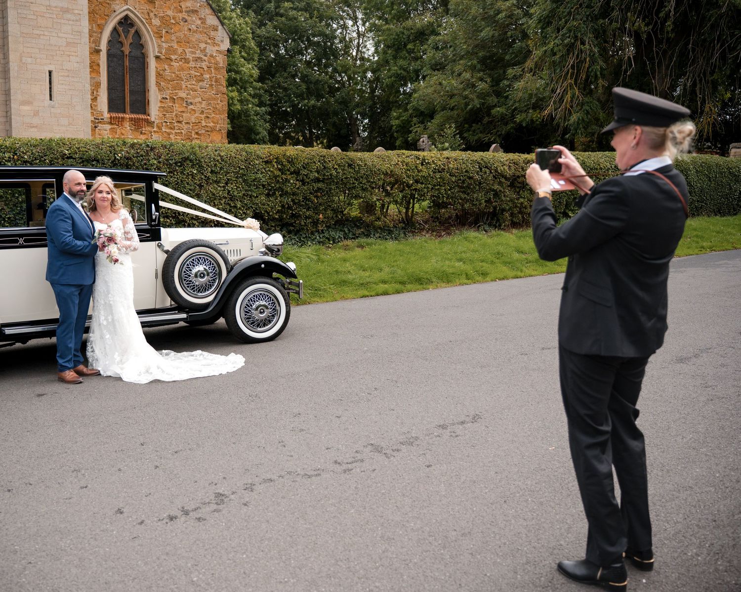 Chauffeur photographing bride and groom posing beside a vintage black and white wedding car outside a church.