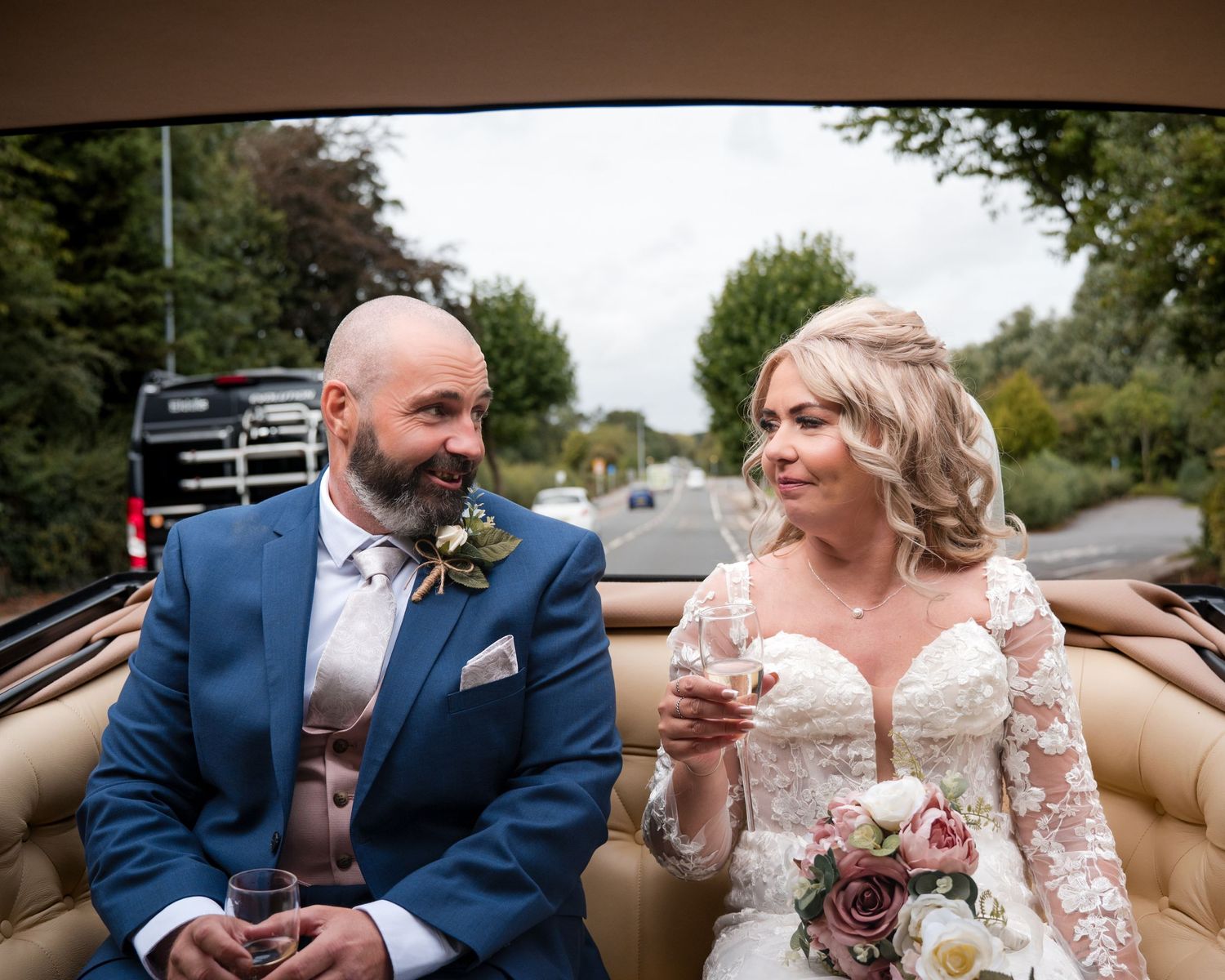 Bride and groom sitting in a vintage convertible car, smiling at each other on their wedding day.