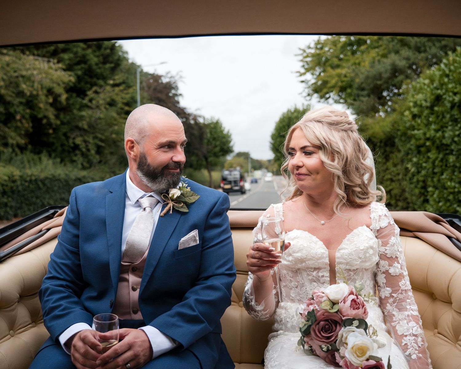Bride and groom riding in an open-top vintage car, smiling and holding champagne glasses on their wedding day.